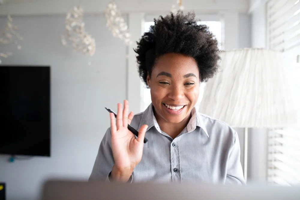 A woman with short curly hair smiling and waving while sitting in front of a laptop in a bright room.