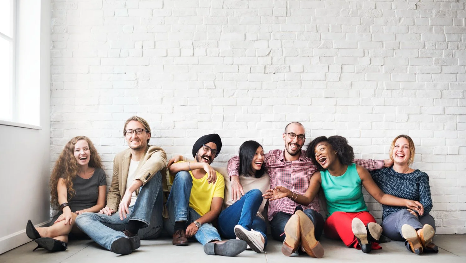 Group of seven diverse people sitting on the floor, smiling and laughing against a white brick wall in a bright room.