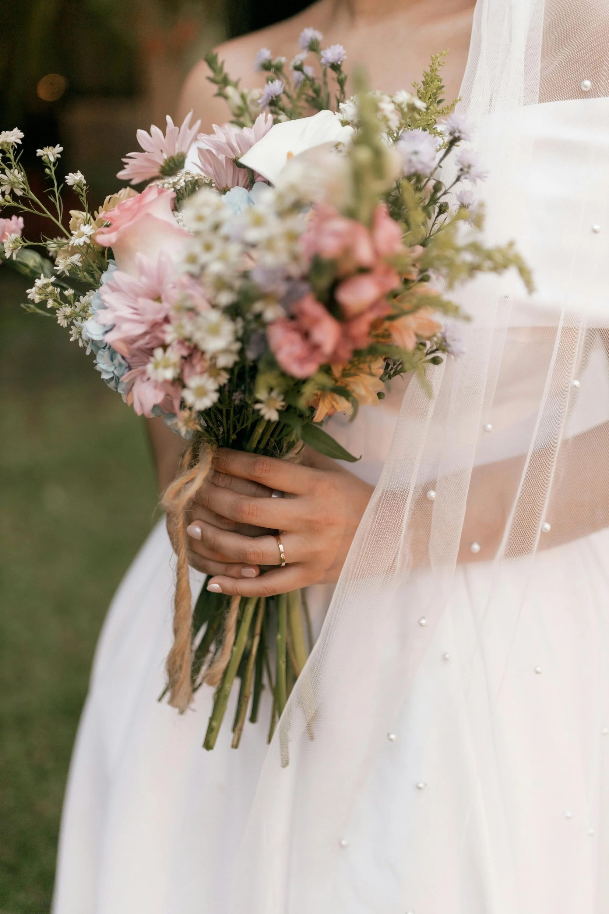 Close-up of a bride holding a bouquet of pink, white, and purple flowers, wearing a wedding dress with pearl embellishments and a sheer veil.