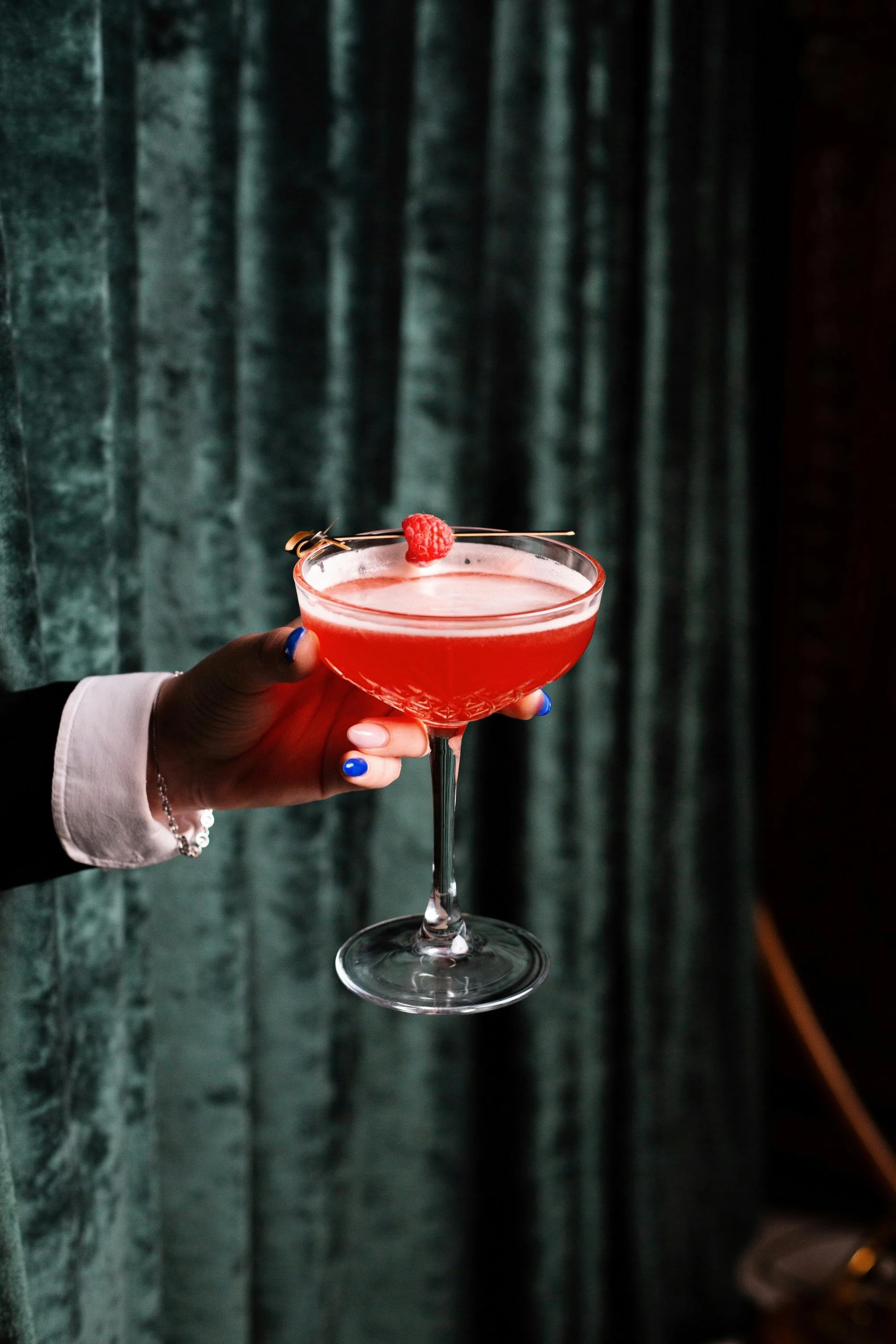 A person holding a pink cocktail garnished with a raspberry in front of a dark green curtain.