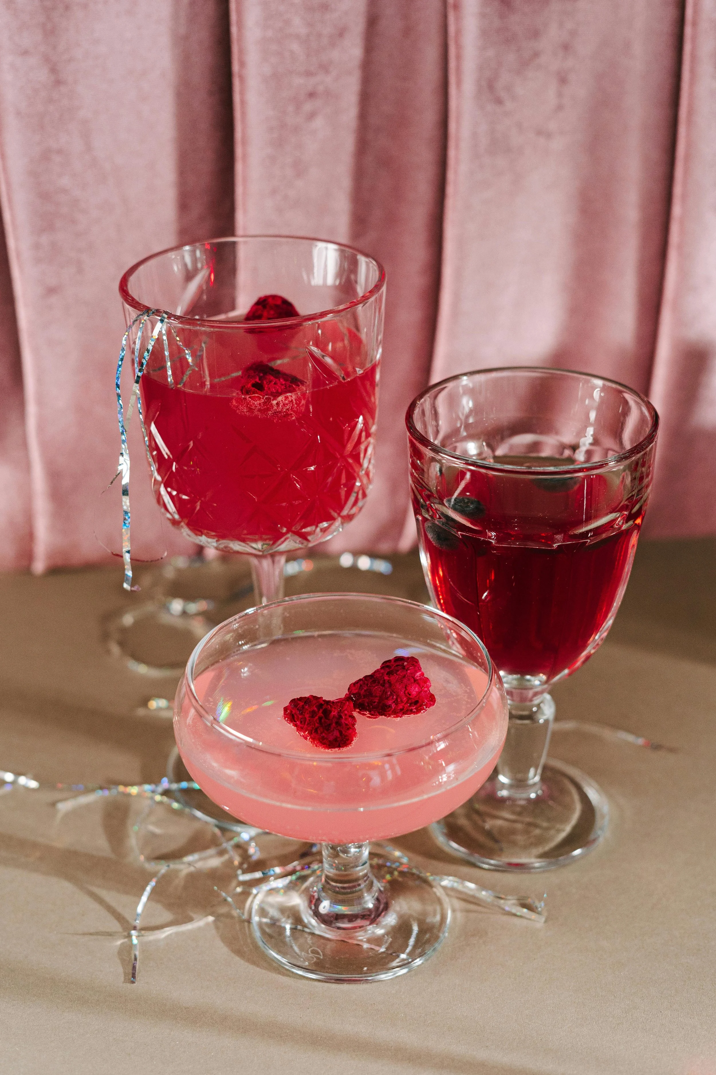 Three glasses with red and pink beverages, garnished with raspberries, on a table with pink curtains in the background and some decorative silver ribbons.