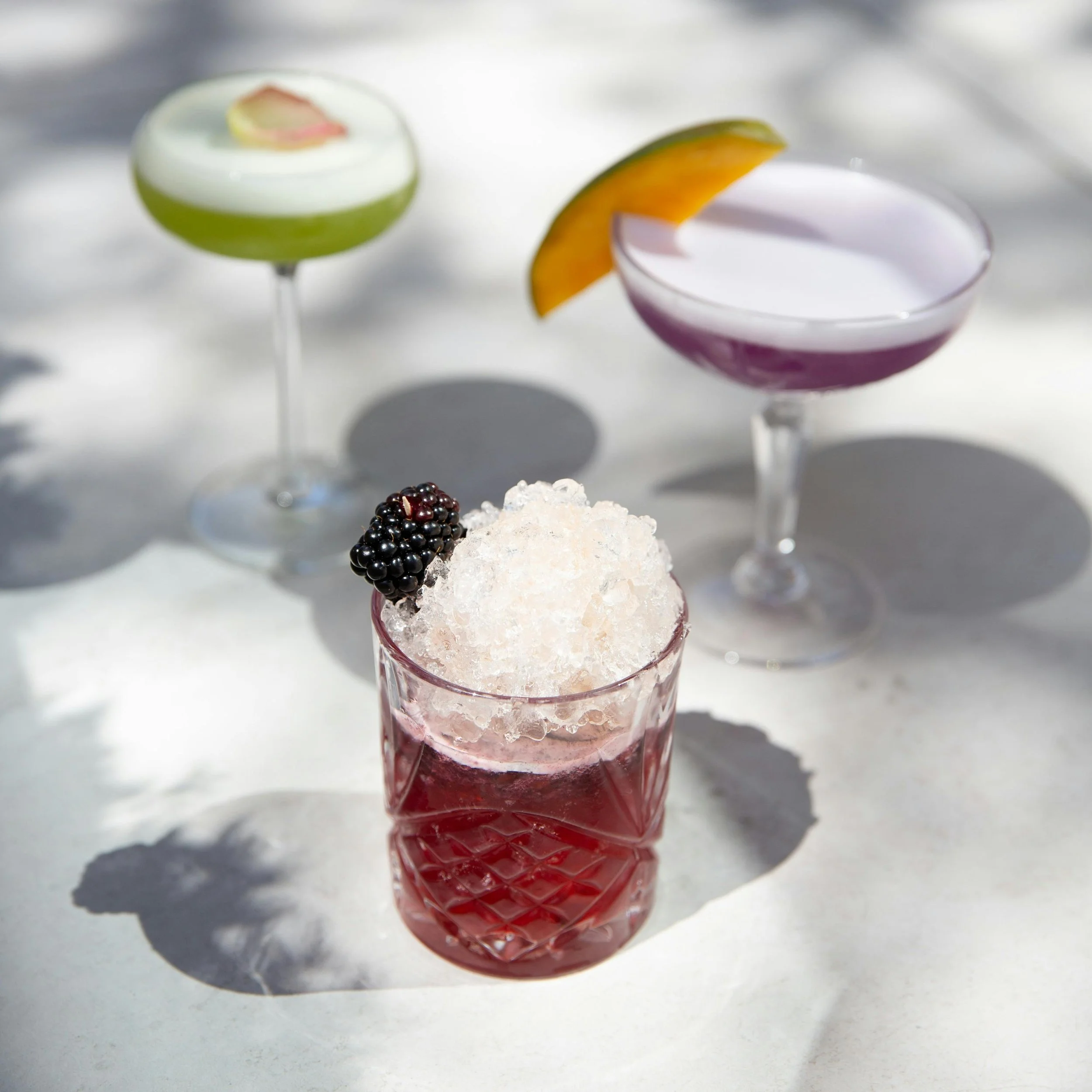 Three colorful cocktails on a white surface, with one garnished with blackberries and crushed ice, and the others with fruit slices.