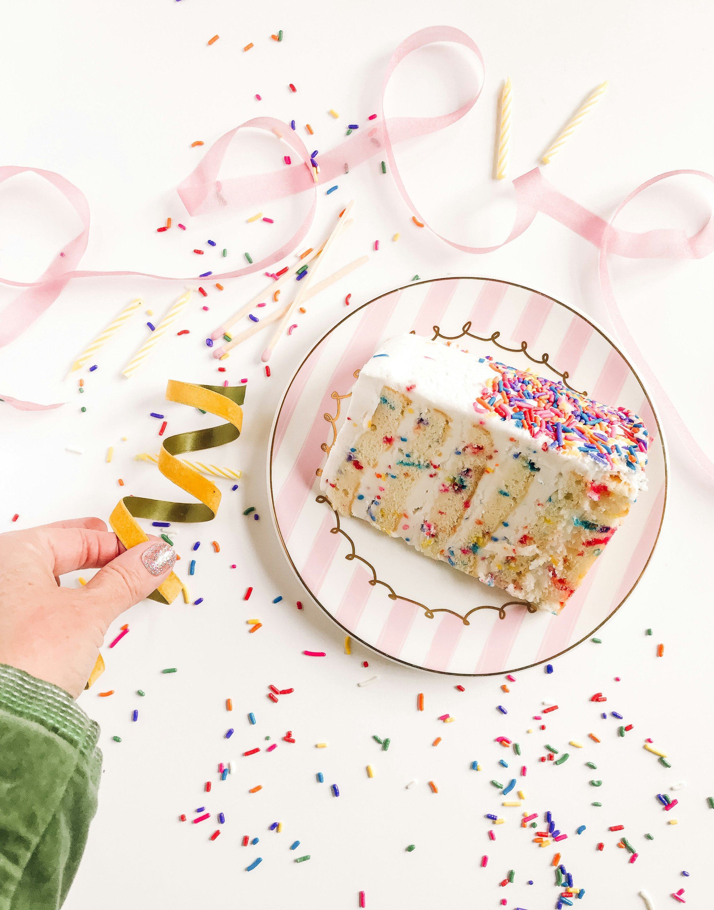 A birthday cake with white frosting, colorful rainbow sprinkles on top, served on a pink and white striped plate. Scattered multicolored confetti, pink ribbon, and unlit birthday candles surround the cake on a white surface. A hand holding a gold and yellow curled ribbon is in the bottom left corner.