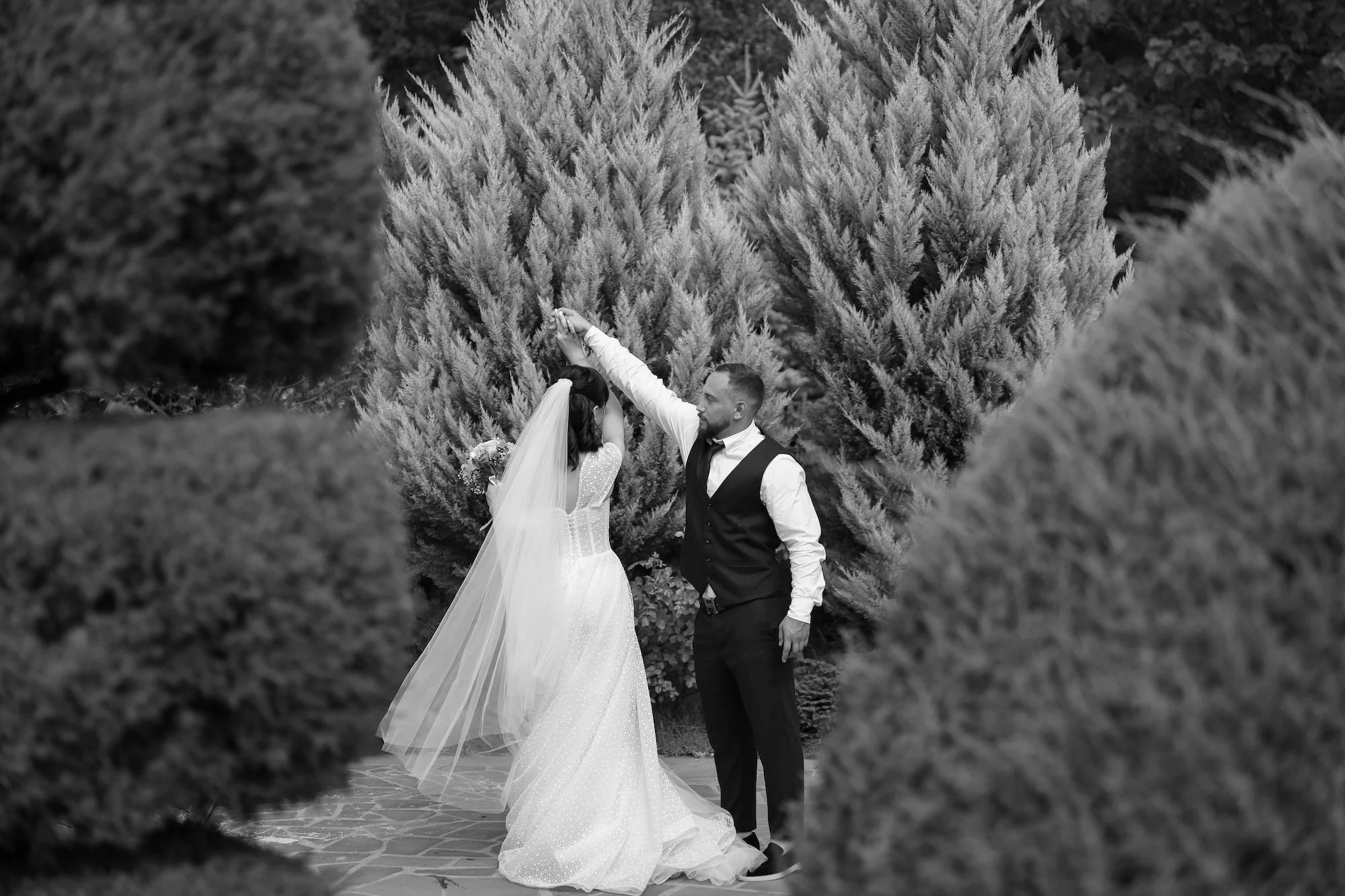 A bride and groom dancing outdoors, surrounded by large bushes and trees, with the groom raising his arm.