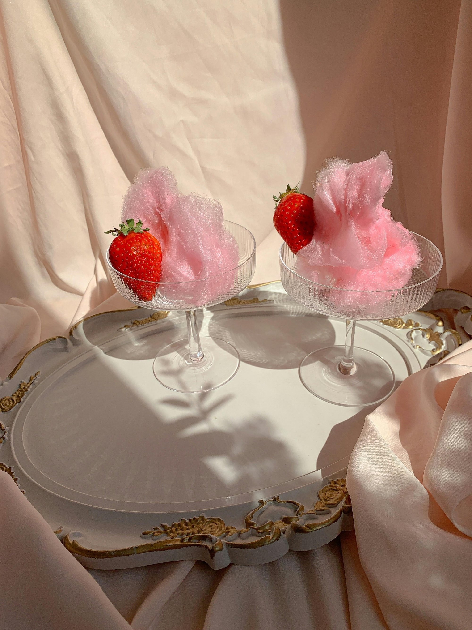 Two cocktail glasses with pink cotton candy and strawberries, placed on a decorative tray with draped fabric and light background.