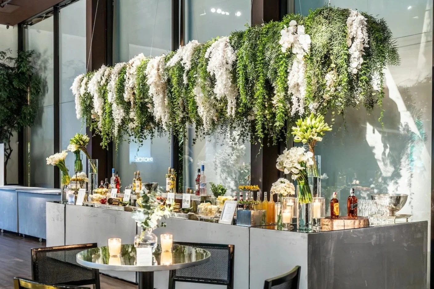 A bar setup decorated with an abundance of white flowers and green foliage, with bottles of alcohol and drink garnishes behind the counter, and lit candles on a round table with a floral centerpiece in the foreground.