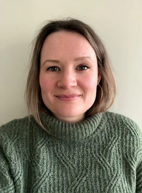 Pictured is a white lady with brown shoulder-length hair and a green jumper. She is smiling.