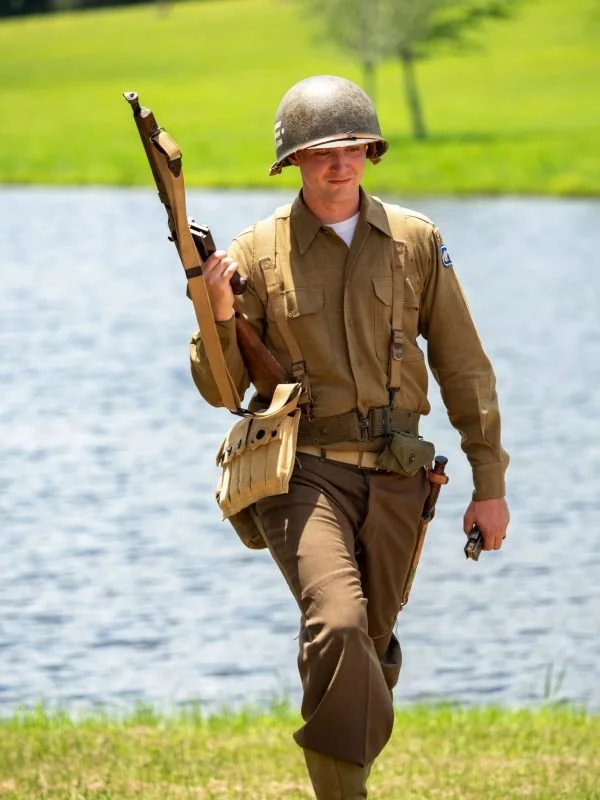 A soldier in uniform walking near a lake, holding a rifle in his right hand and a pistol in his left hand, wearing a helmet.