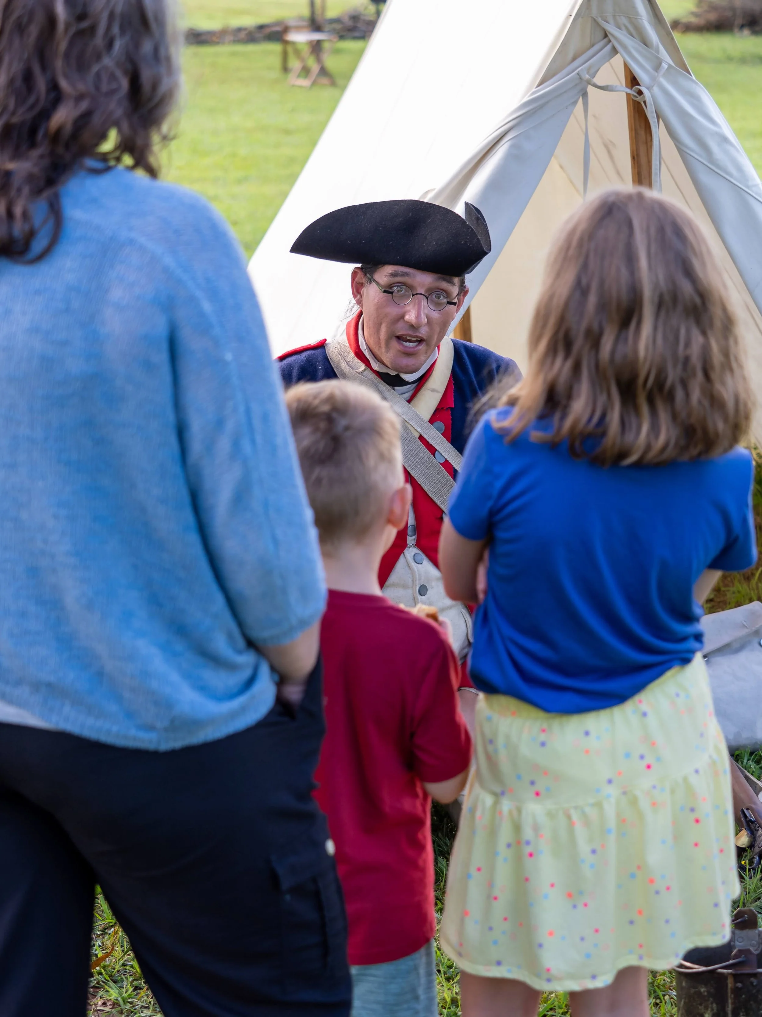 A man dressed as a historical soldier, wearing a tricorn hat and colonial uniform, is talking to three children and a woman near a white tent outdoors, with green grass and trees in the background.