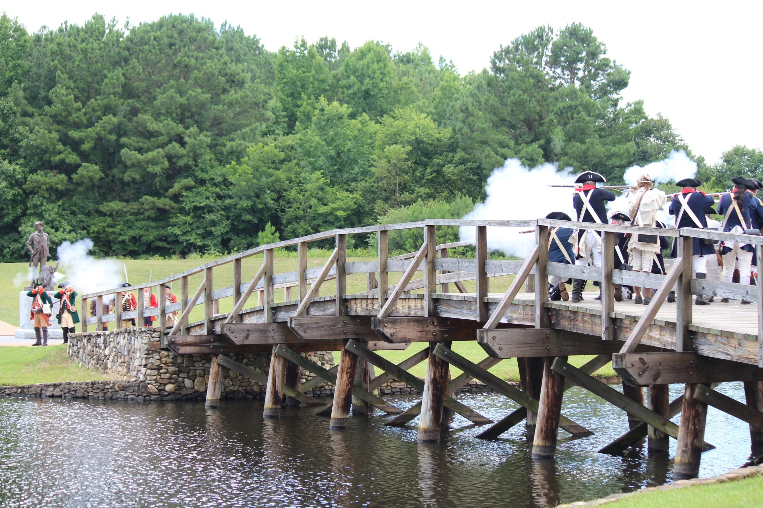 Historical reenactment of a battle with soldiers firing muskets on a wooden bridge over water, surrounded by green trees.
