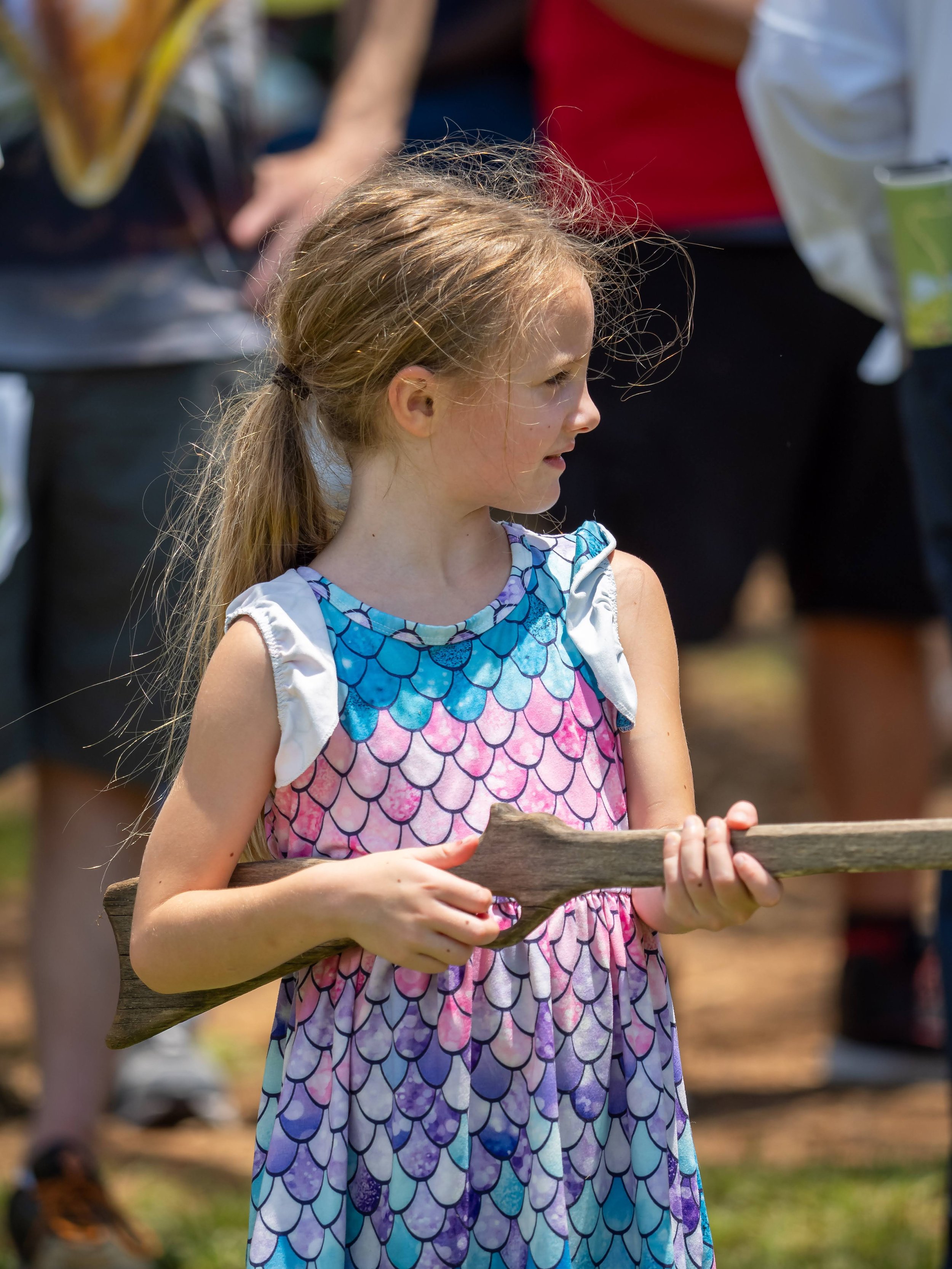 A young girl with long hair tied in a ponytail, holding a wooden stick, wearing a colorful dress with a fish scale pattern, outdoors at a sunny event with people in the background.