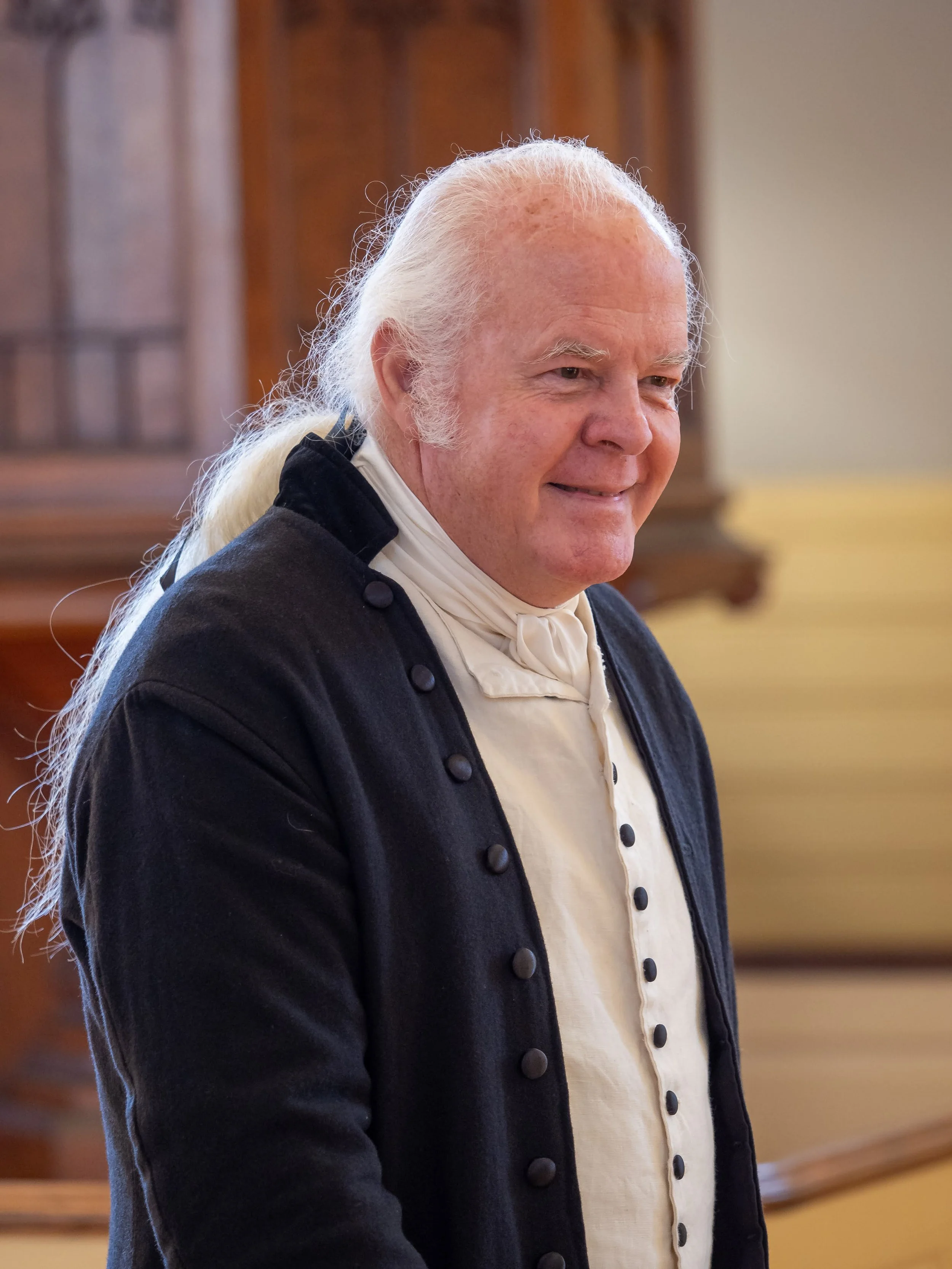 A smiling elderly man with long white hair tied back, dressed in historical attire with a black coat and white shirt, inside a wood-paneled room.