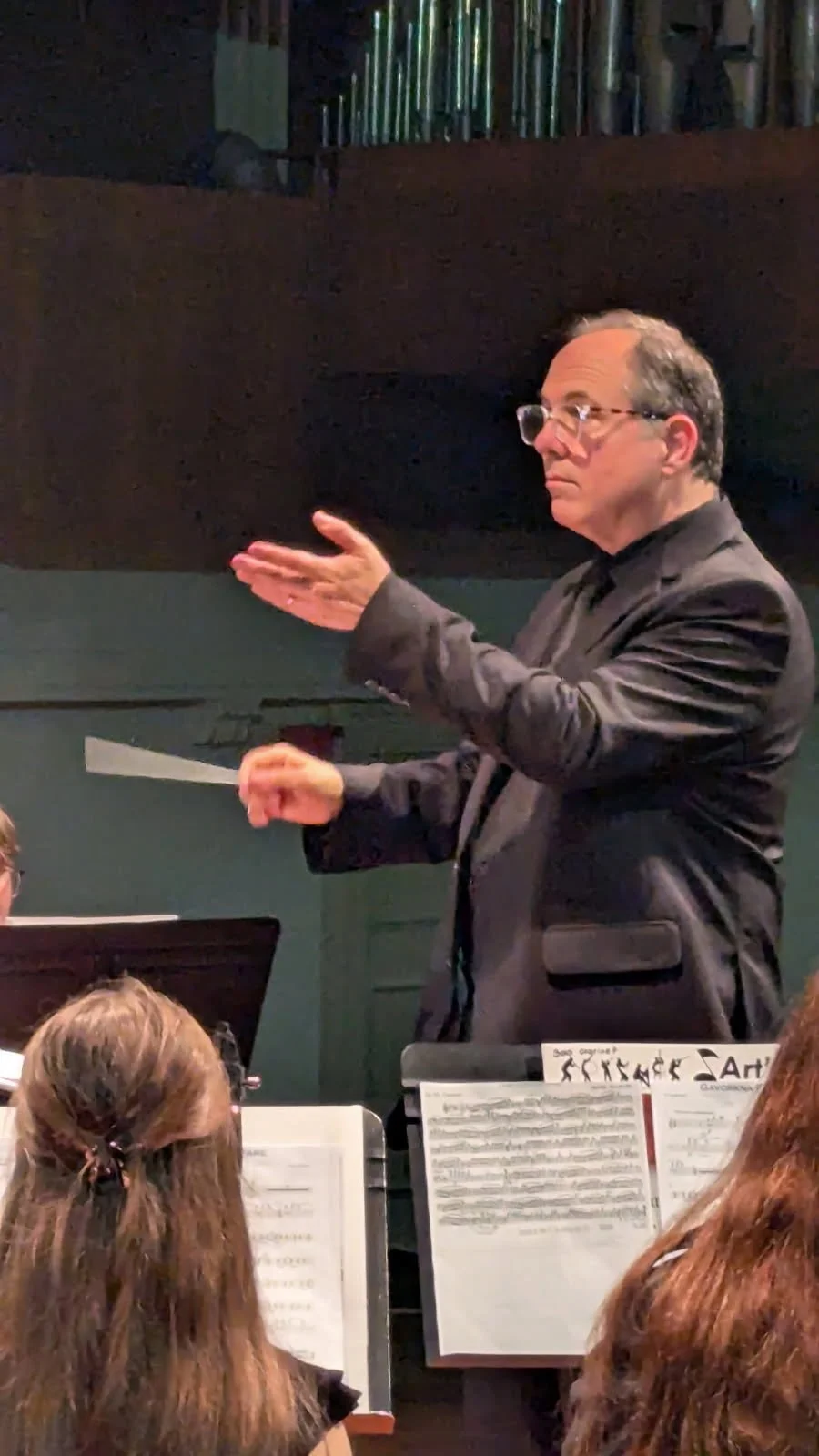 Joe Ardovino wearing glasses and a dark suit conducts an orchestra or choir during a performance, with sheet music visible in front of him.