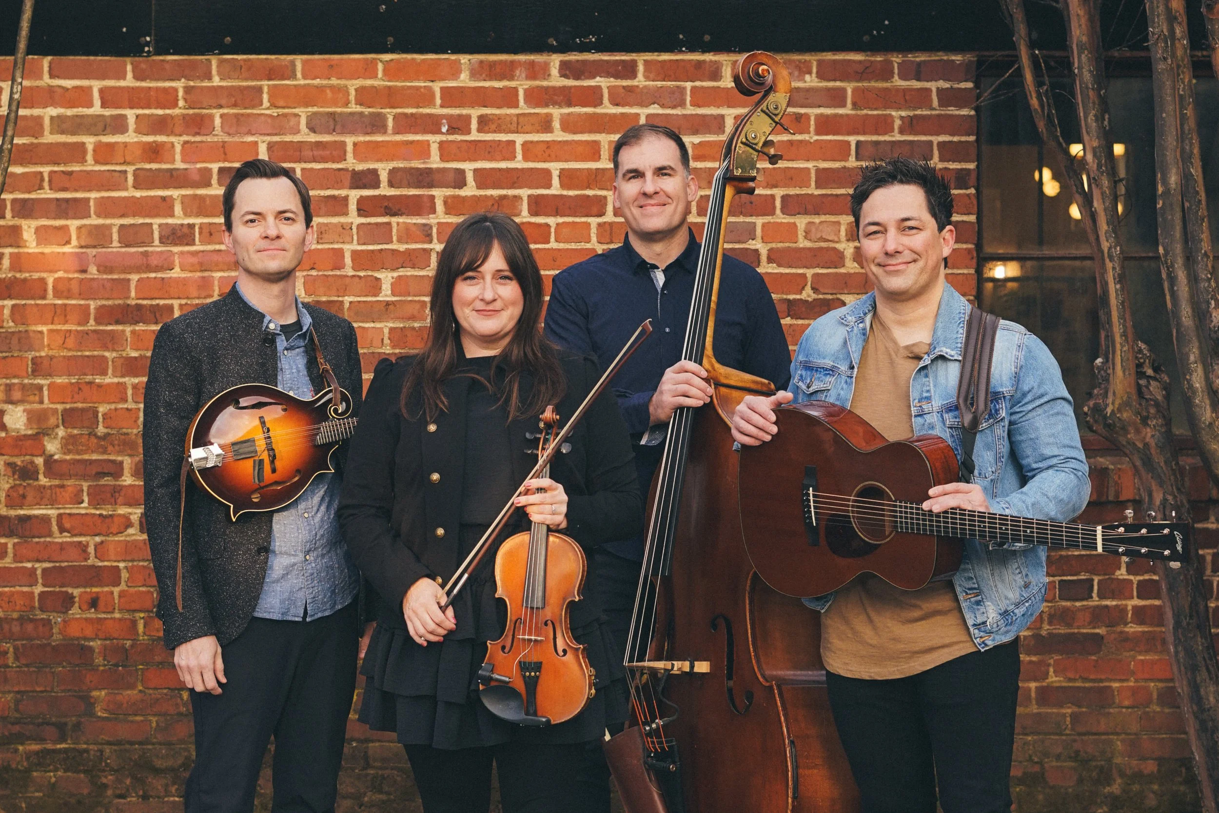 Act of Congress standing in front of a brick wall, each holding a musical instrument: a mandolin, a violin, a double bass, and a guitar, smiling for the camera.