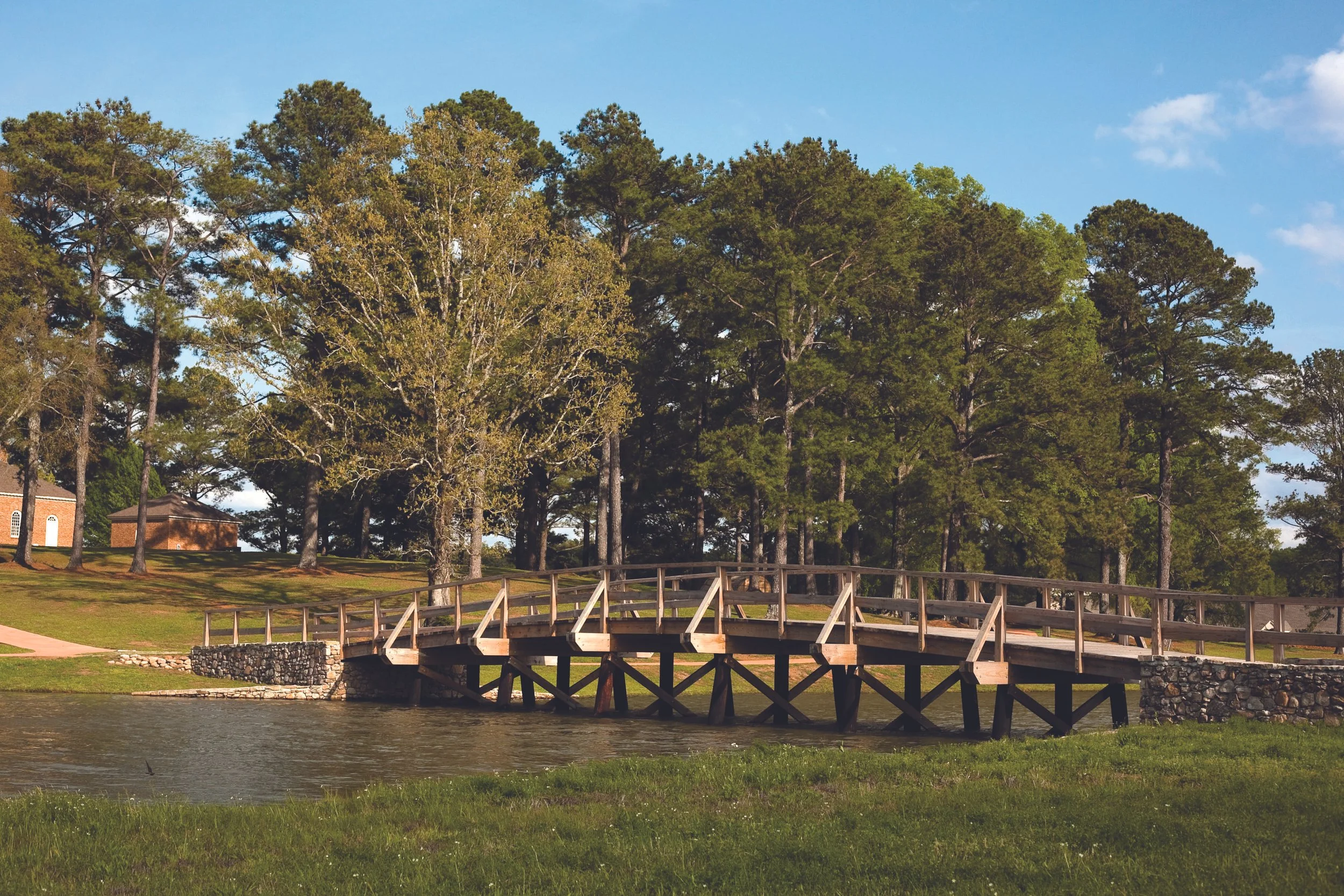 A wooden bridge over a small river, surrounded by lush green grass and large trees, under a partly cloudy blue sky.