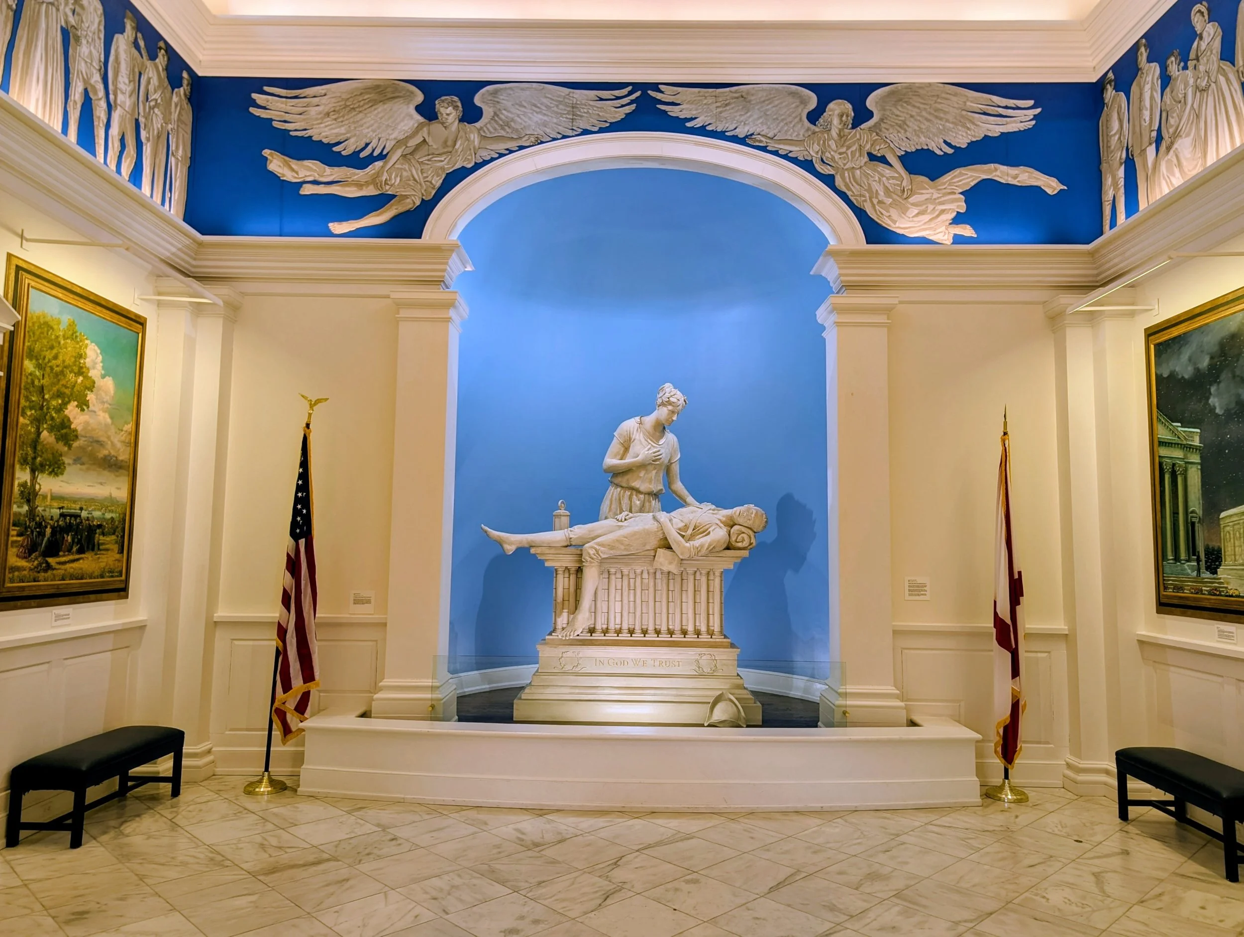 A museum exhibit featuring a white marble sculpture of a woman kneeling beside a lifeless man, with American flags on either side. The wall behind is painted blue, with white relief sculptures of angels and figures.