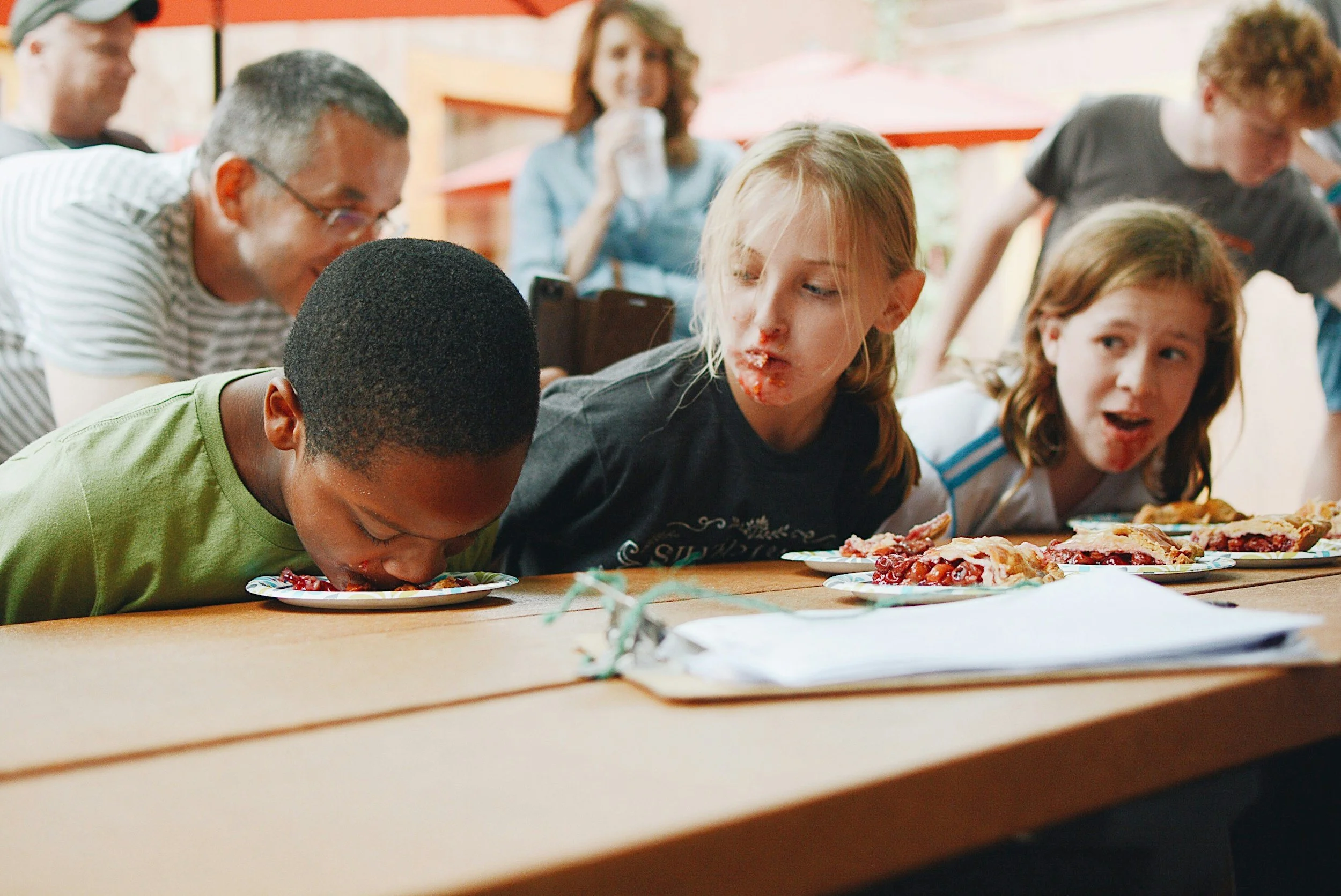 Young children at a pizza-eating contest with plates of pizza in front of them, some with food on their faces, and adults in the background watching.