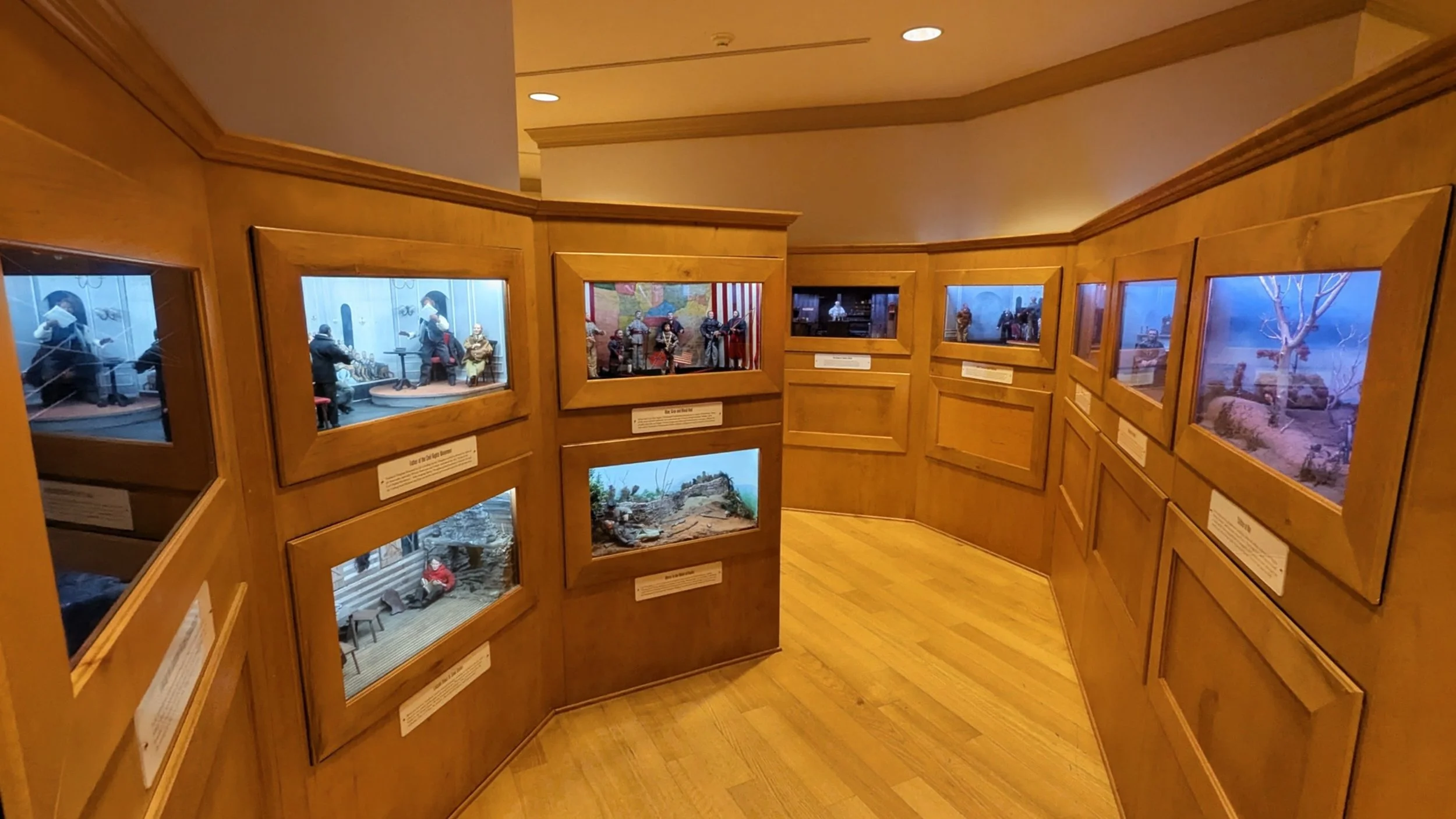 Exhibit display with multiple dioramas and miniature scenes in a room with wooden paneling and flooring.