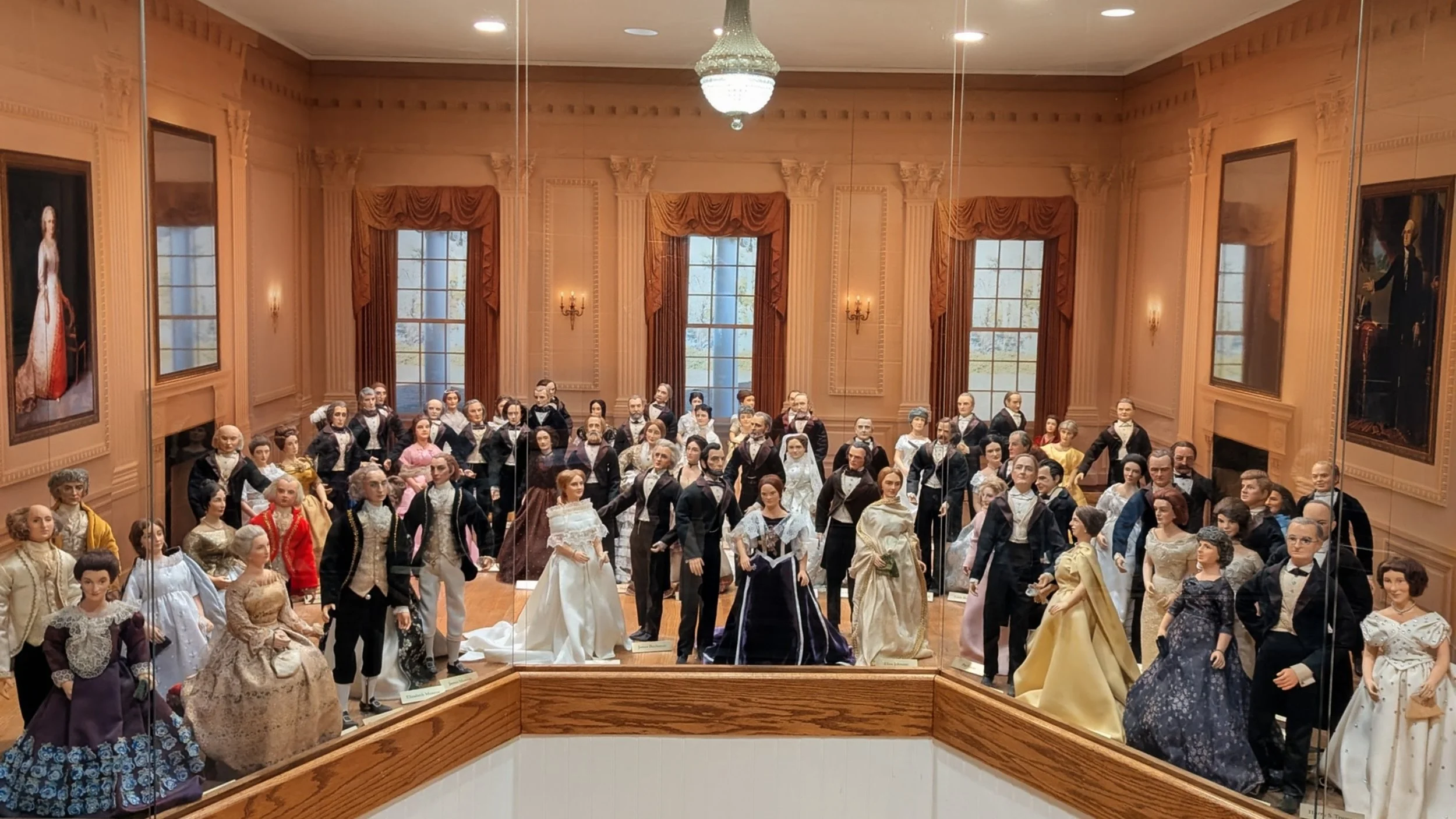 Glass display case containing a large collection of wax figures dressed in 19th-century attire, arranged in a room with wood-paneled walls, large windows with red curtains, and portrait paintings.