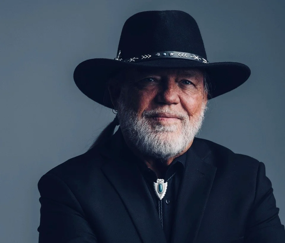 Rick Carter wearing a black cowboy hat with decorative band, black jacket, and bolo tie, smiling against a neutral background.