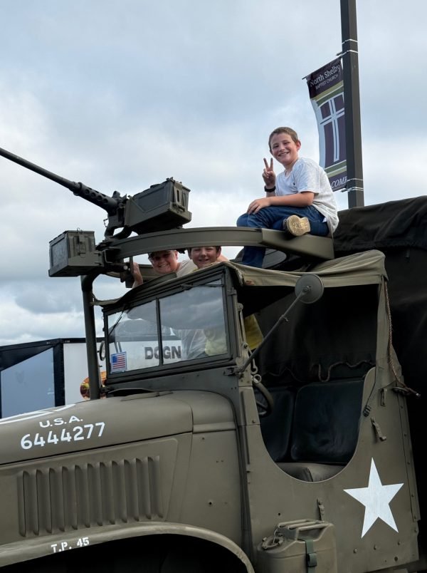 Three children sitting on top of a military vehicle, with one making a peace sign and all smiling, outdoors on a cloudy day.