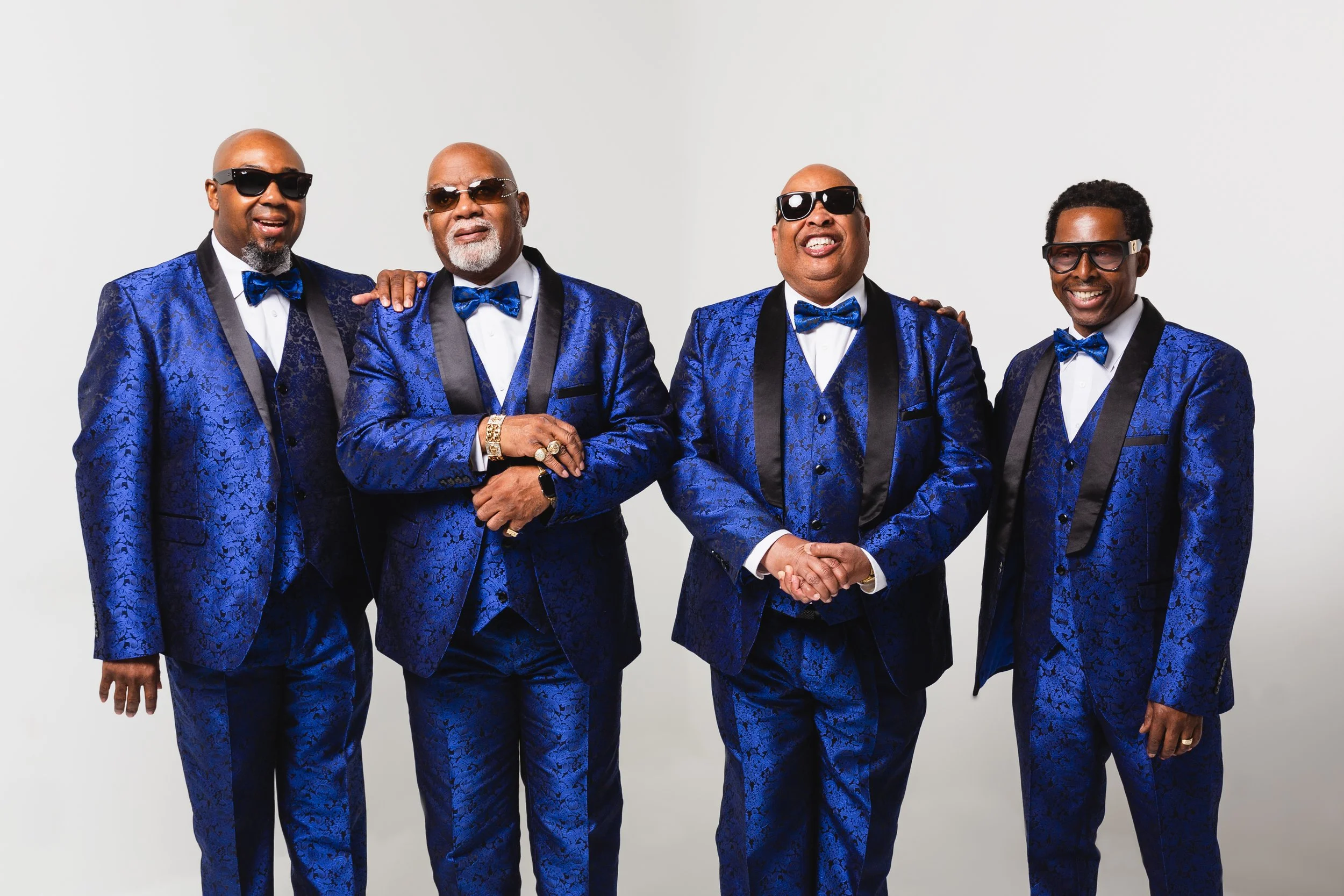 The Blind Boys of Alabama dressed in matching blue tuxedos with black lapels, bow ties, and sunglasses, standing together against a white background. The men are smiling and one is holding hands with the man next to him.
