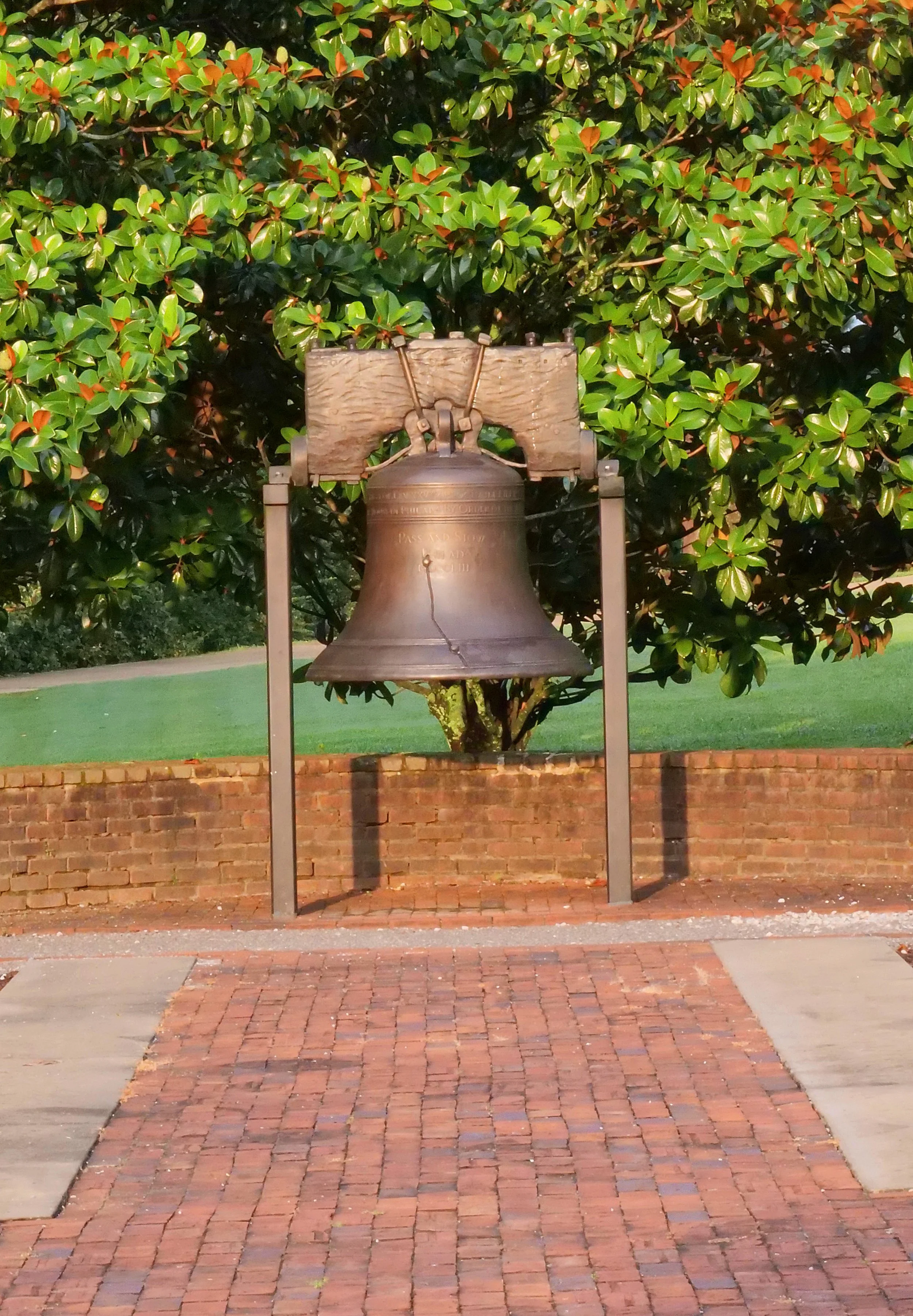 A large bronze bell hanging in a wooden frame, situated outdoors in front of a green leafy tree and a brick wall pathway.