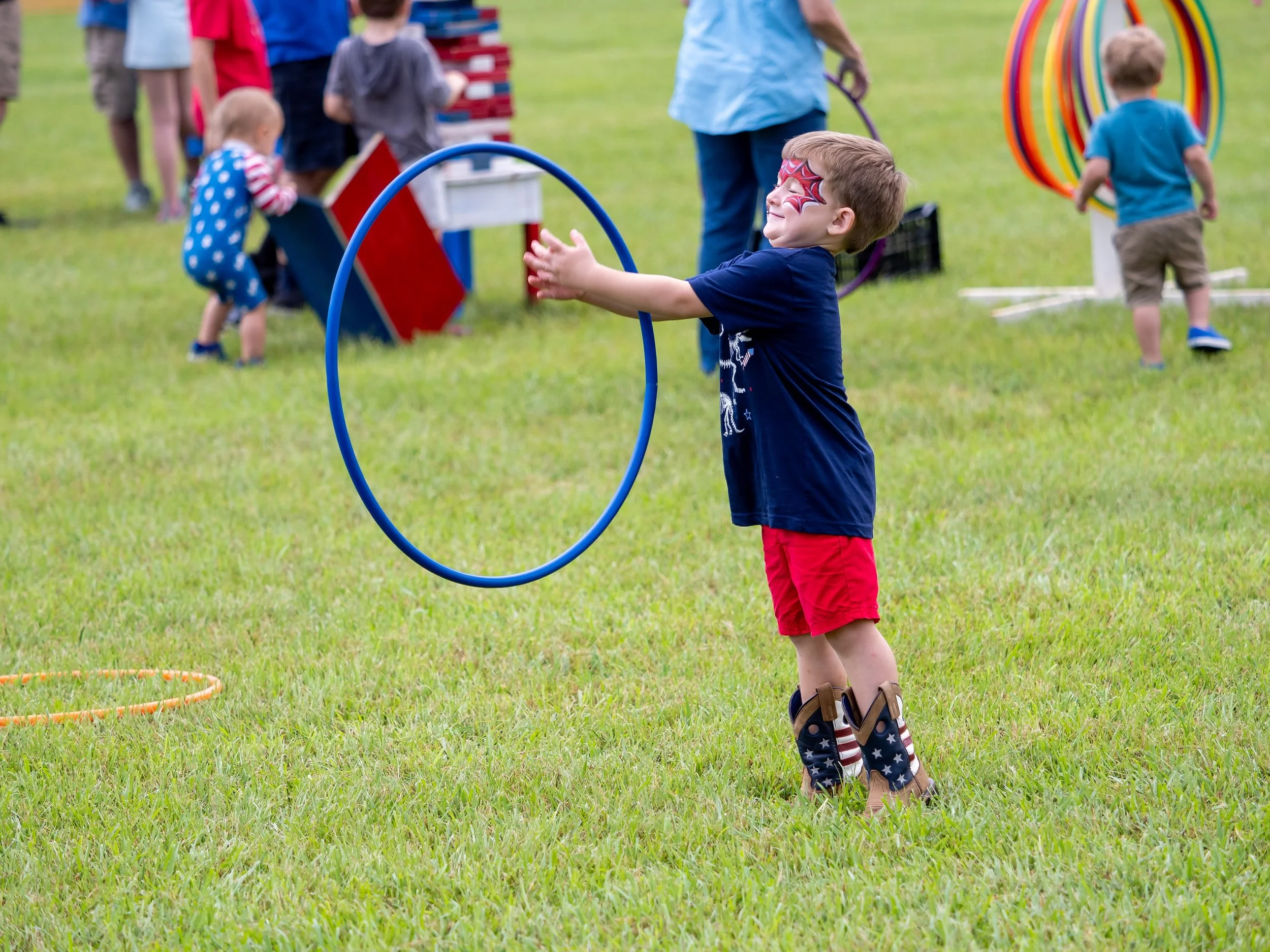 A young boy in red shorts and cowboy boots playing with a hula hoop at an outdoor event, with children and adults in the background engaging in activities on a grassy field.