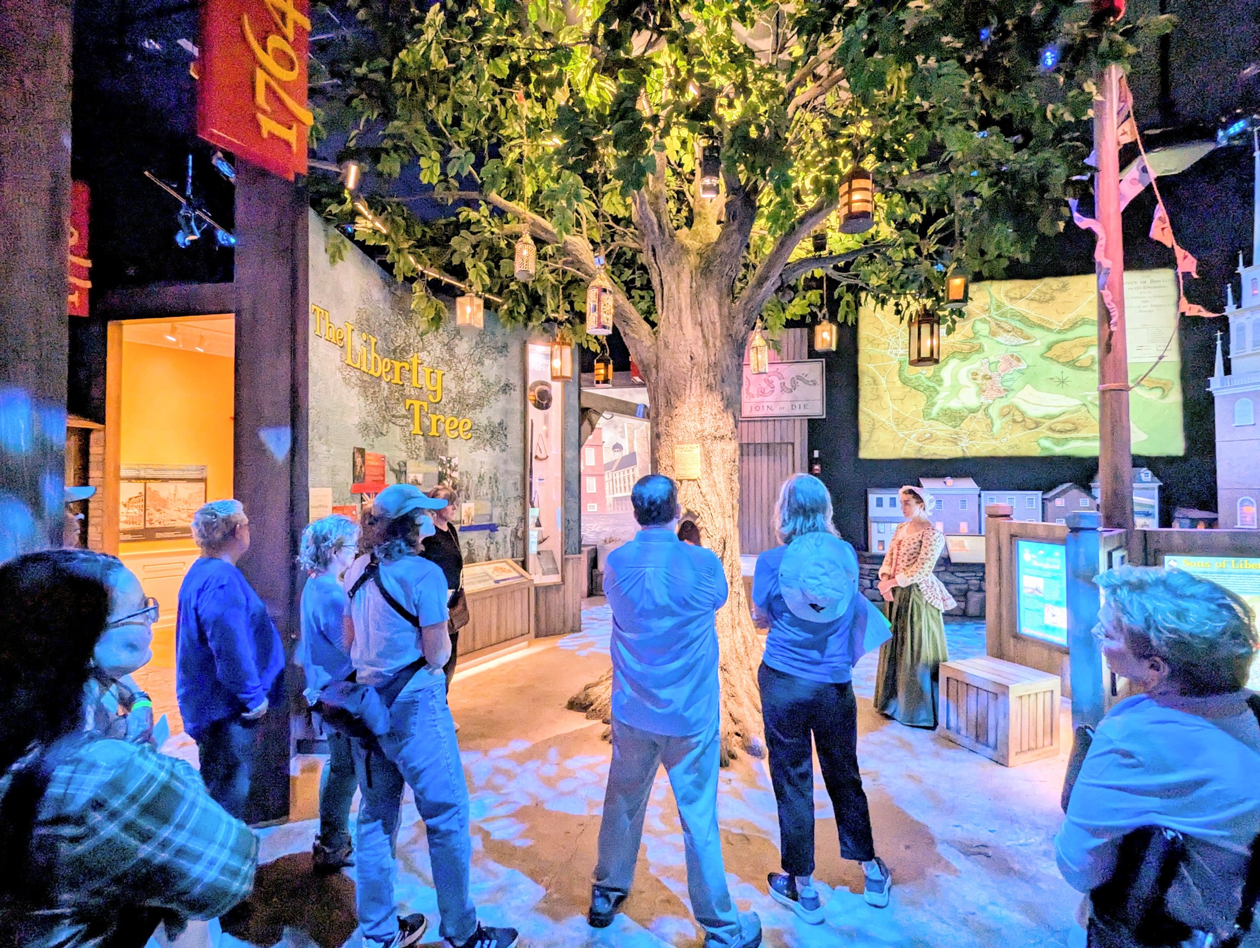 Group of people gathered around a storytelling exhibit called 'The Liberty Tree' featuring a large tree and a historical theme.