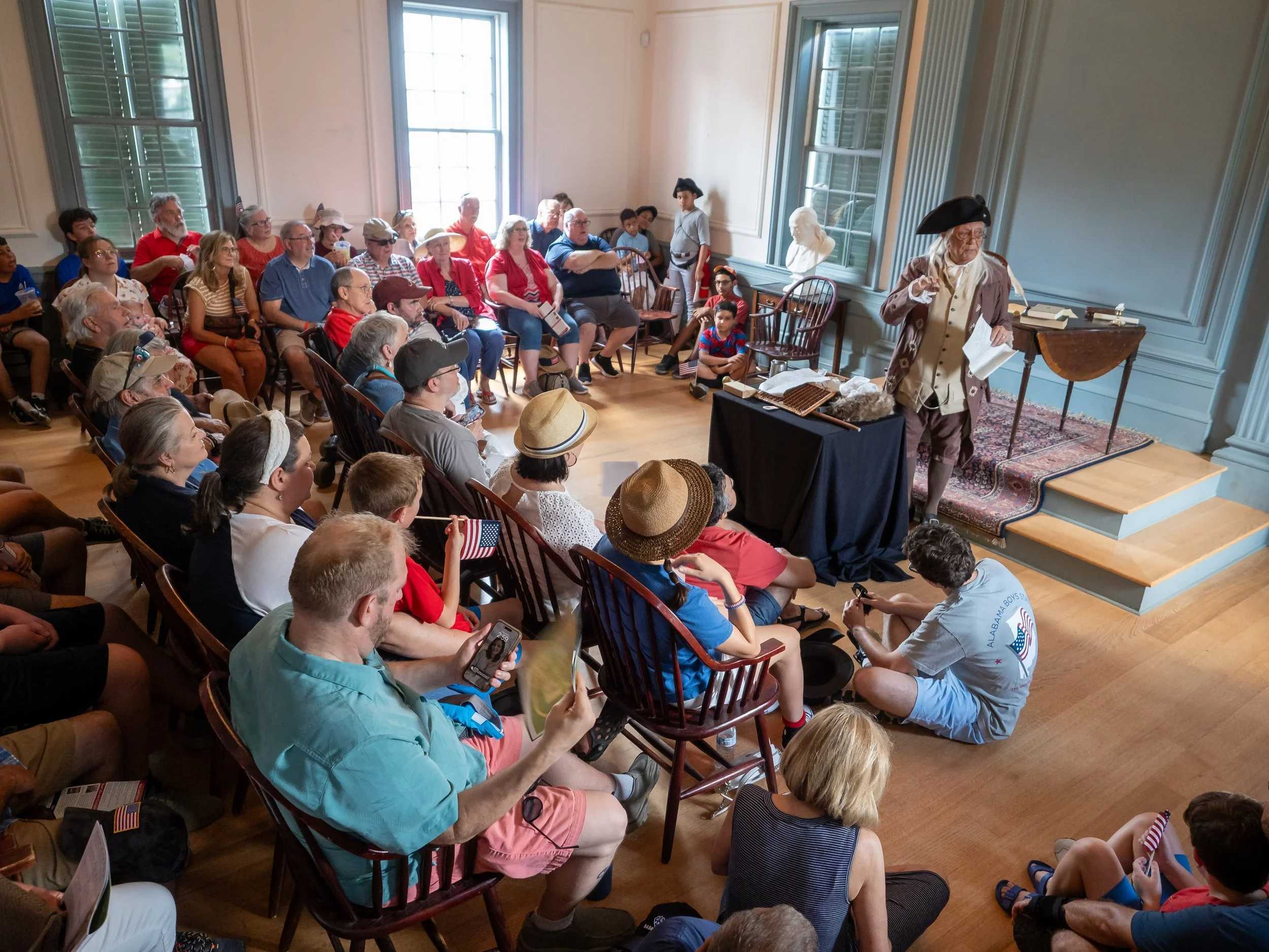 A historical reenactment in a museum featuring a man dressed as a colonial figure speaking to a seated audience of various ages, with some audience members holding small American flags.