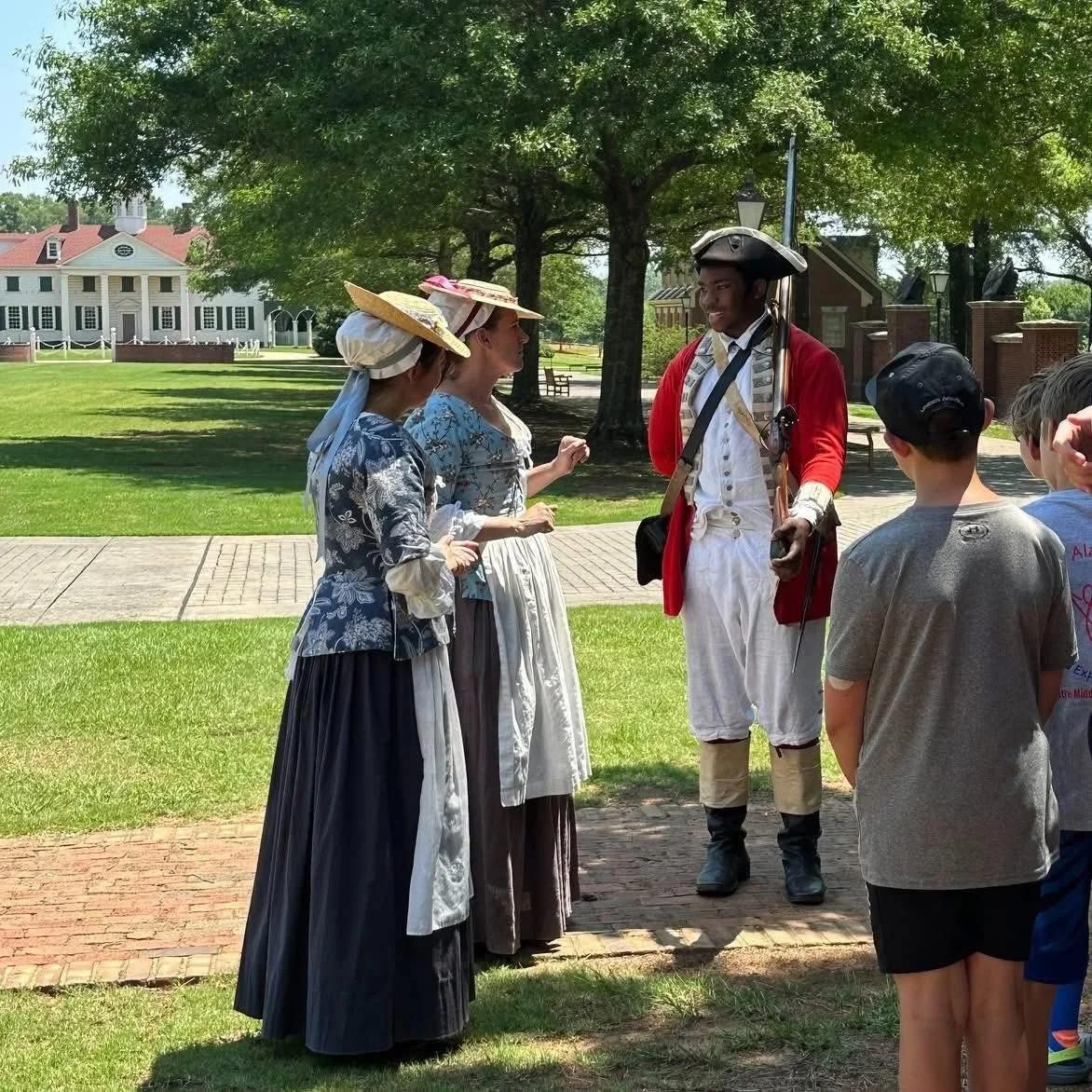 People dressed in colonial-era clothing, including tricorn hats and coats, standing outdoors on a sunny day with trees and a historic building in the background.