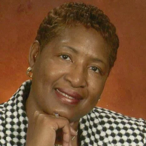 Peggy Towns with short curly hair, wearing a checkered blazer, smiling, with a warm background.