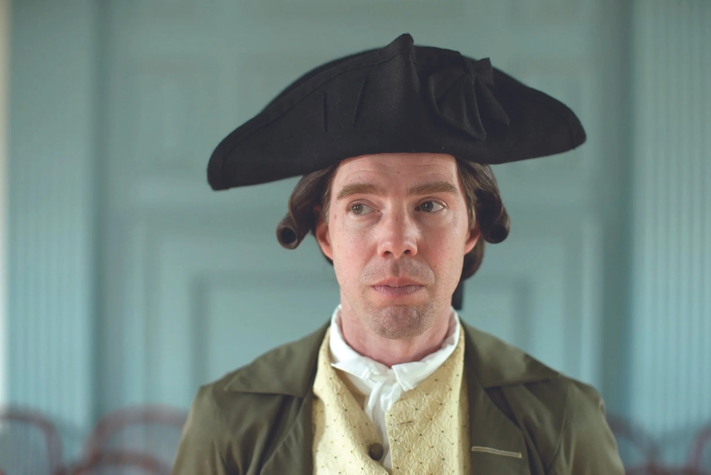 Man dressed in 18th-century costume wearing a black tricorn hat, cream-colored waistcoat, and olive green coat, with a neutral background.