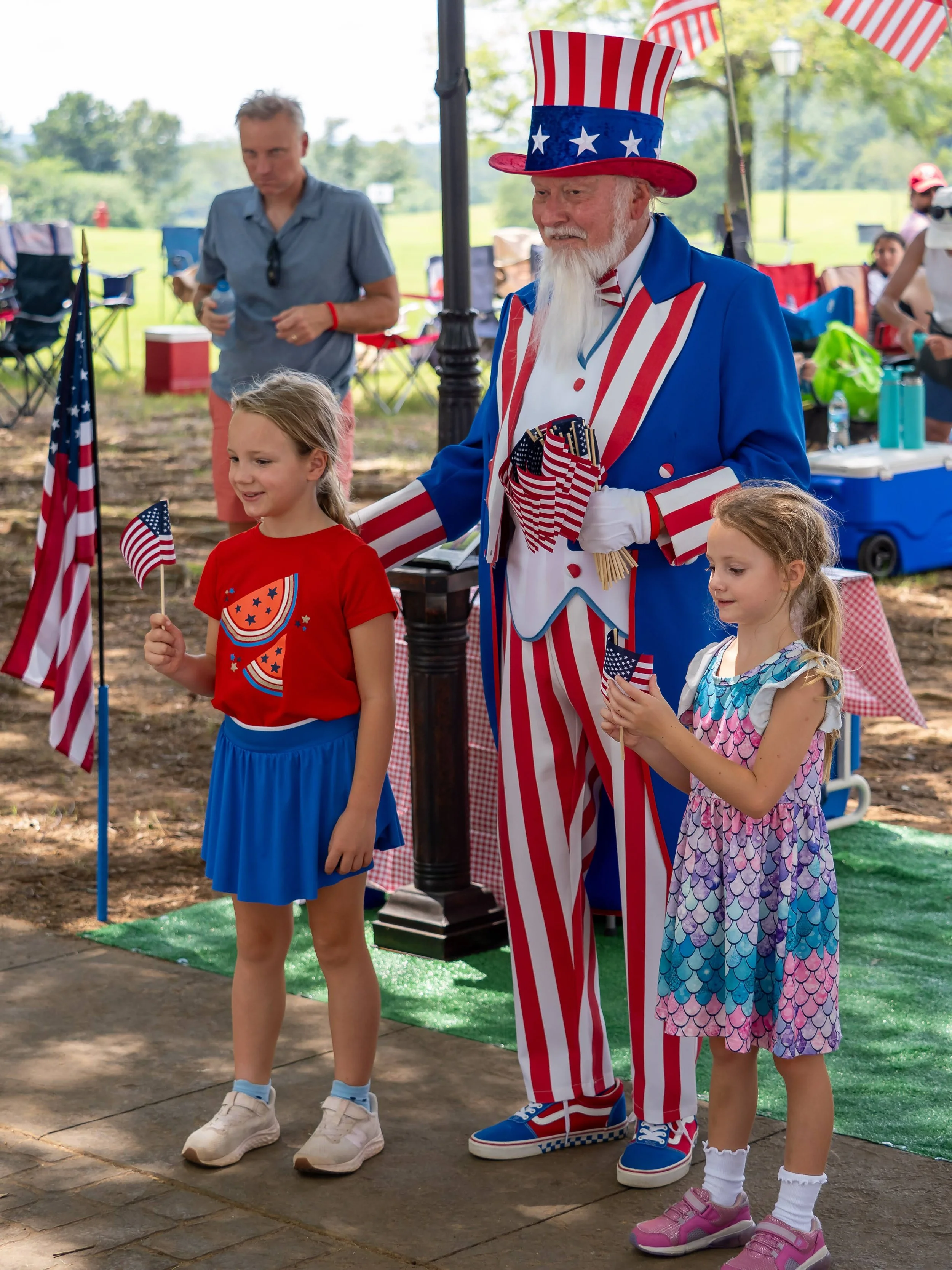 Two young girls standing next to a man dressed as Uncle Sam at an outdoor patriotic celebration, holding small American flags. The girls are smiling, and the setting is a park with picnic tables and people in the background.