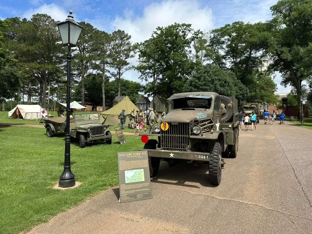 A military exhibit with vintage trucks and cars, tents, and soldiers, set on a grassy park with trees.