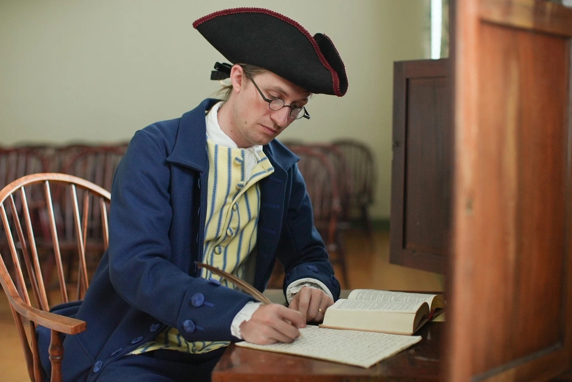 A man dressed in 18th-century colonial attire, wearing a tricorn hat, glasses, and a blue coat, sitting at a wooden desk and writing in a journal.
