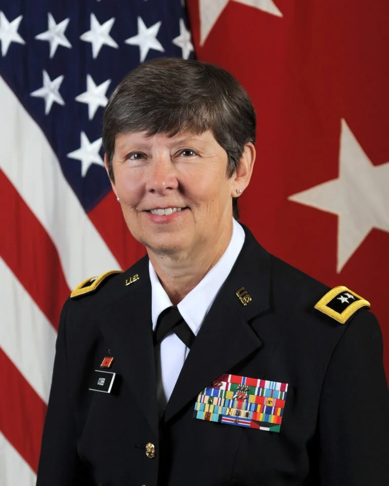 Ret. Maj. Gen. Janet L Cobb in uniform, standing in front of American and red flags, smiling.