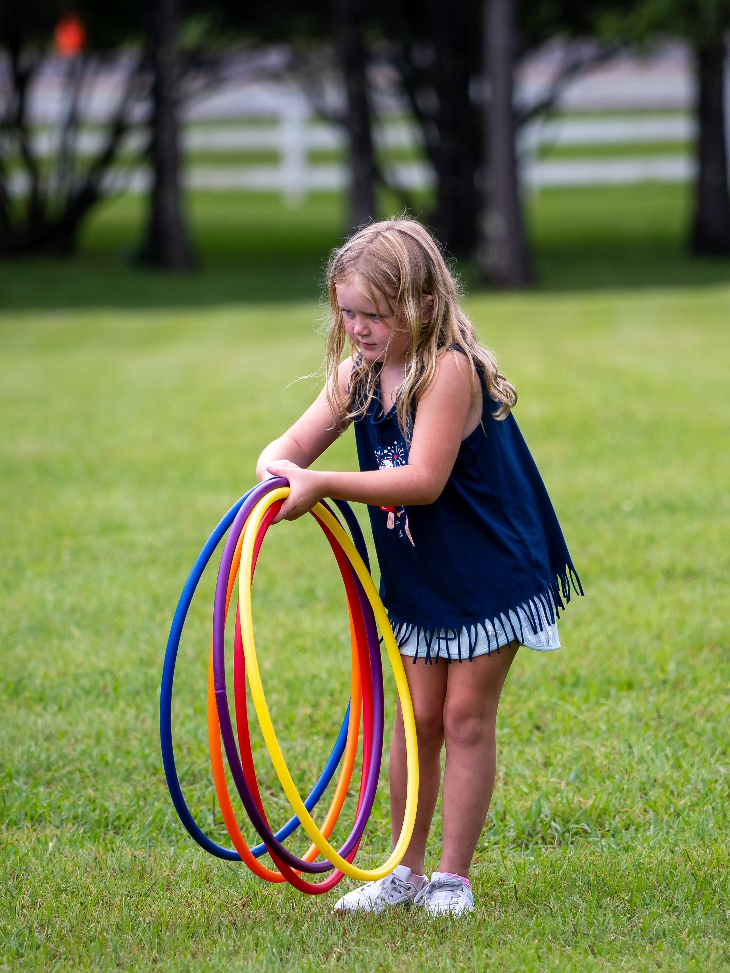 A young girl with long blonde hair, wearing a blue sleeveless top and shorts, is standing on a grassy field holding multiple colorful hula hoops.