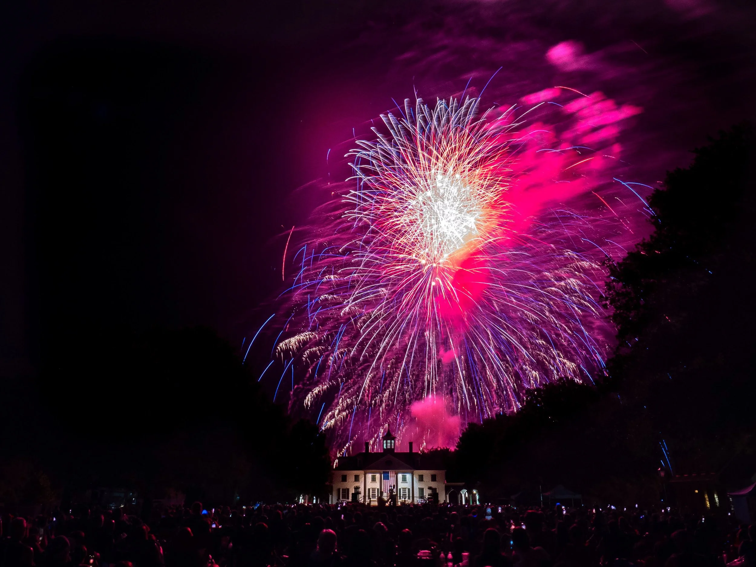 Fireworks in the night sky above a crowd at a celebration, with a large white house illuminated in the foreground.
