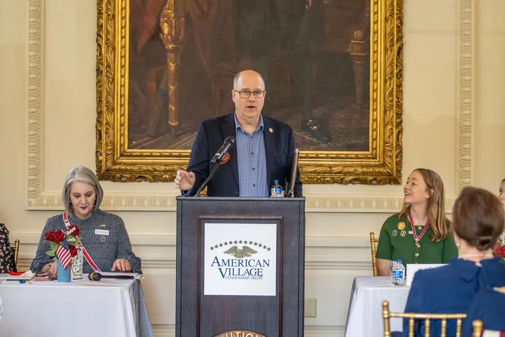 Val Crofts speaking at a podium with the American Village Citizenship Trust logo, flanked by women sitting at tables with patriotic decorations, in a room with ornate gold-framed artwork in the background.