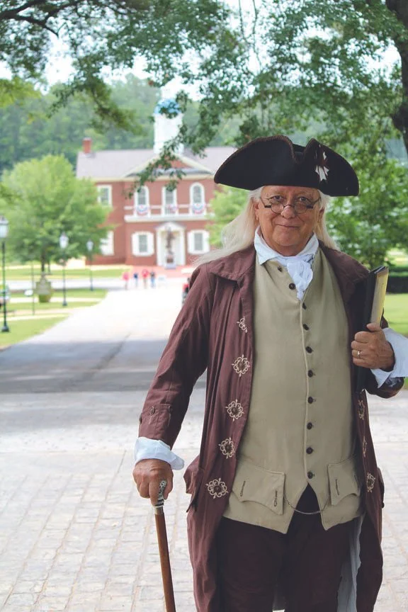 A woman dressed in colonial-era attire, wearing a tricorn hat, glasses, and holding a cane, standing outdoors near a historic building with trees in the background.