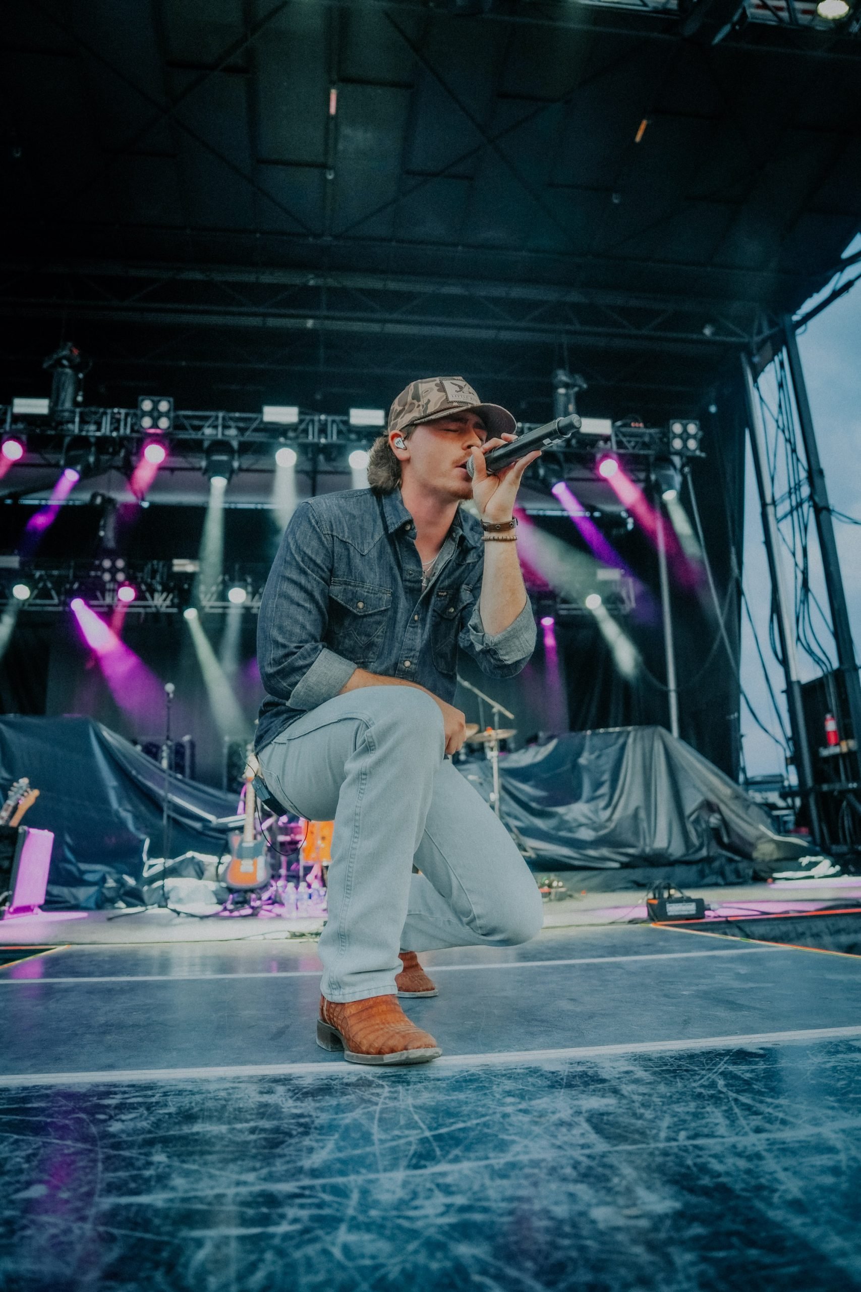 Myles Morgan kneeling on stage with microphone in hand during a concert, stage lights in the background, wearing a cap, denim jacket, jeans, and brown boots.
