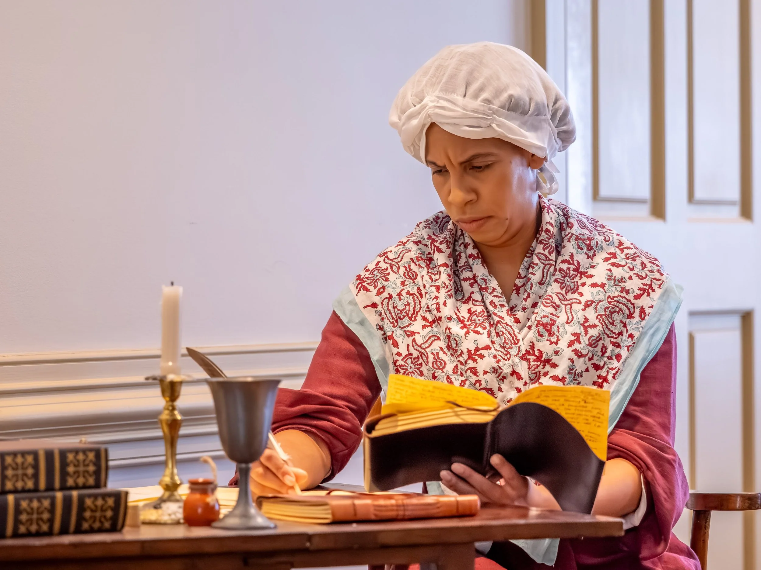 Woman dressed in historical clothing with a bonnet, reading and writing at a table with a candle and old books.