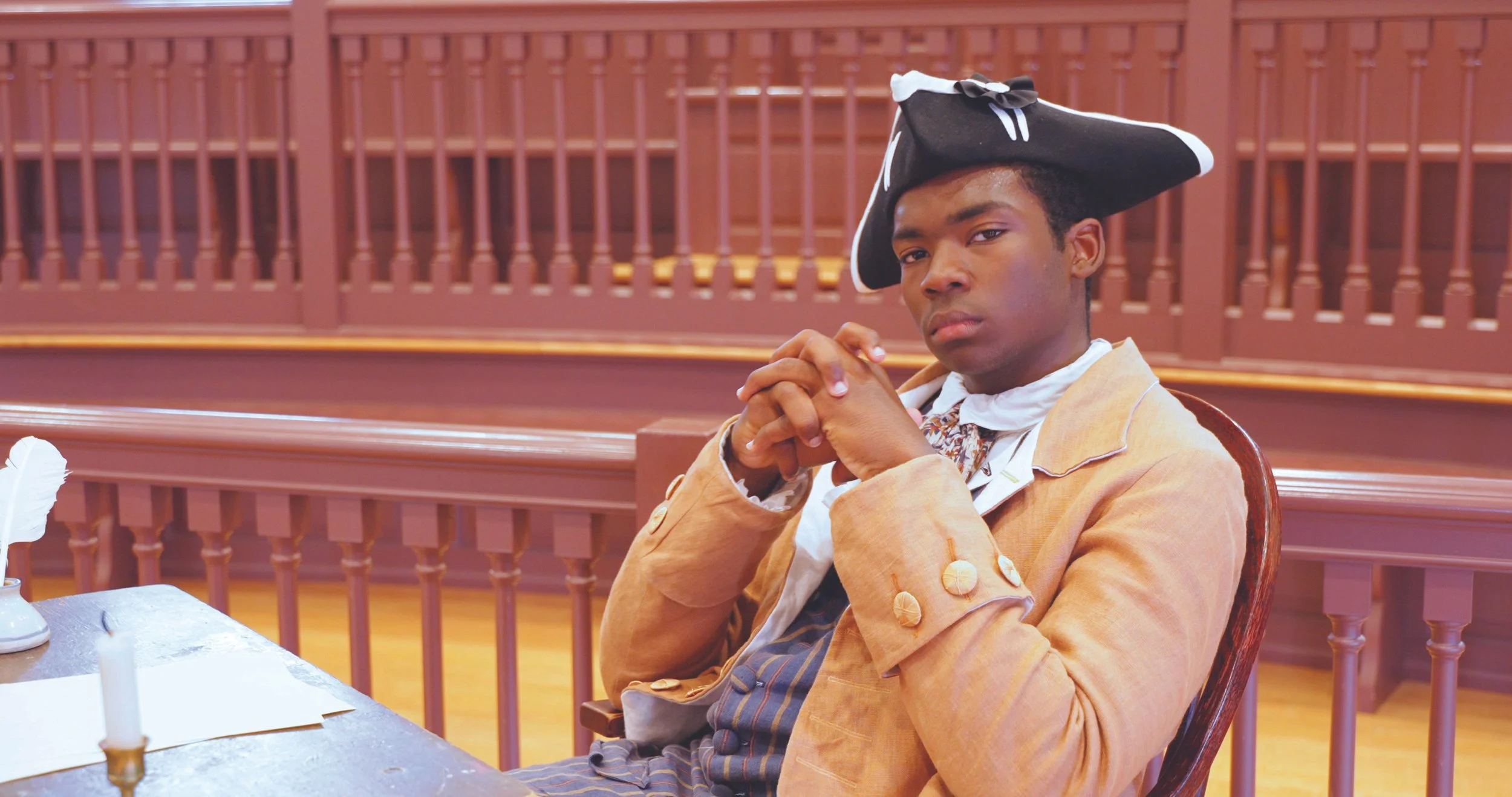 A young man dressed in colonial-era attire with a tricorn hat, sitting at a wooden table in a courtroom or historical setting, with wooden paneling in the background.