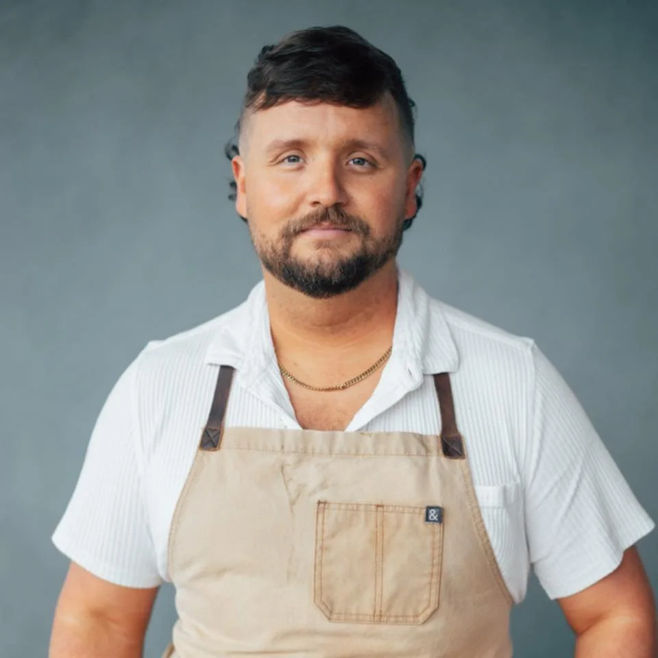 Jonathan Harrison with a beard and short dark hair, wearing a white short-sleeve shirt and a beige apron, standing against a plain gray background.