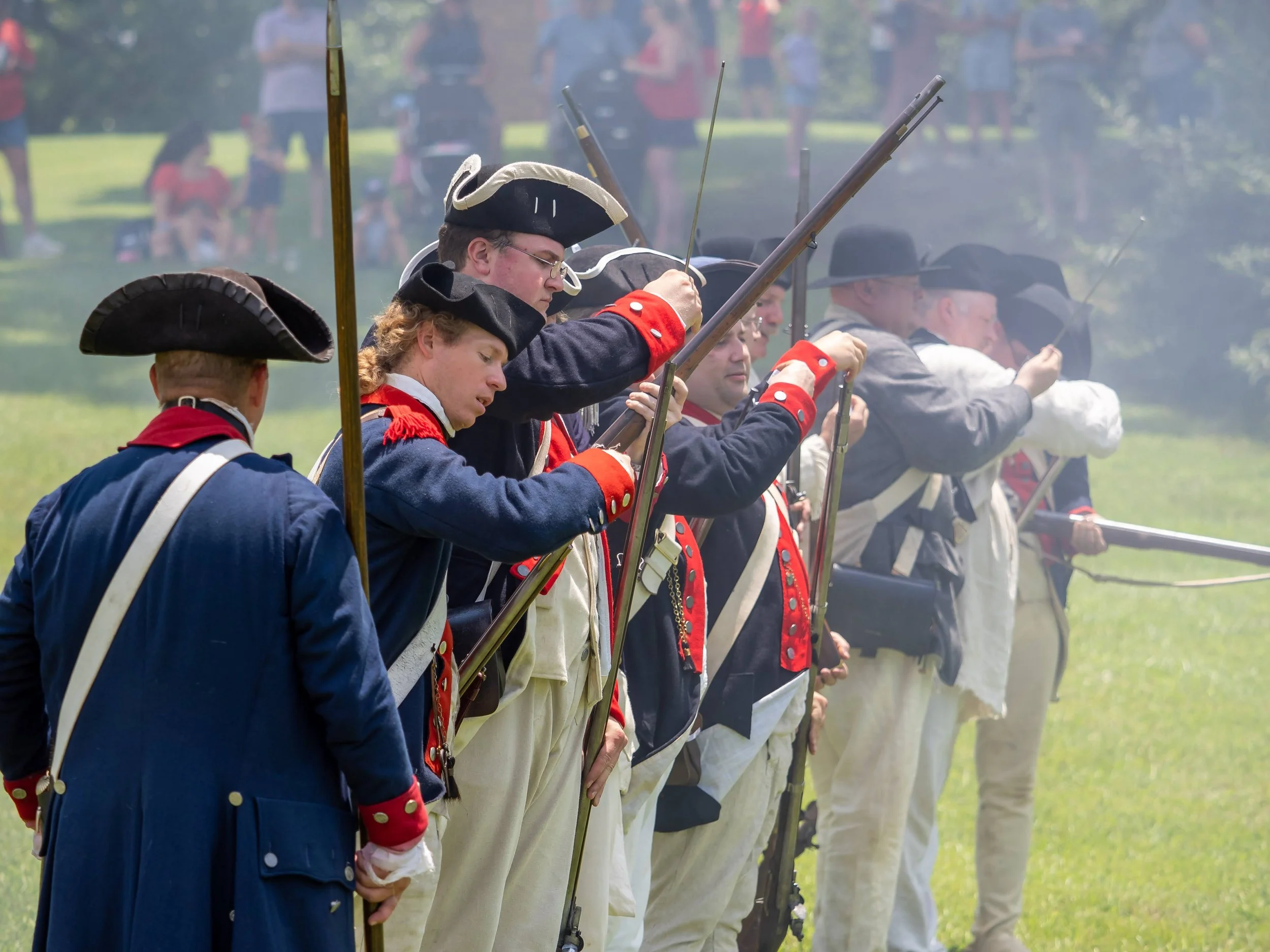 Historical reenactors dressed in 18th-century military uniforms firing muskets or rifles in formation on a grassy field during a reenactment event. Spectators watch in the background.