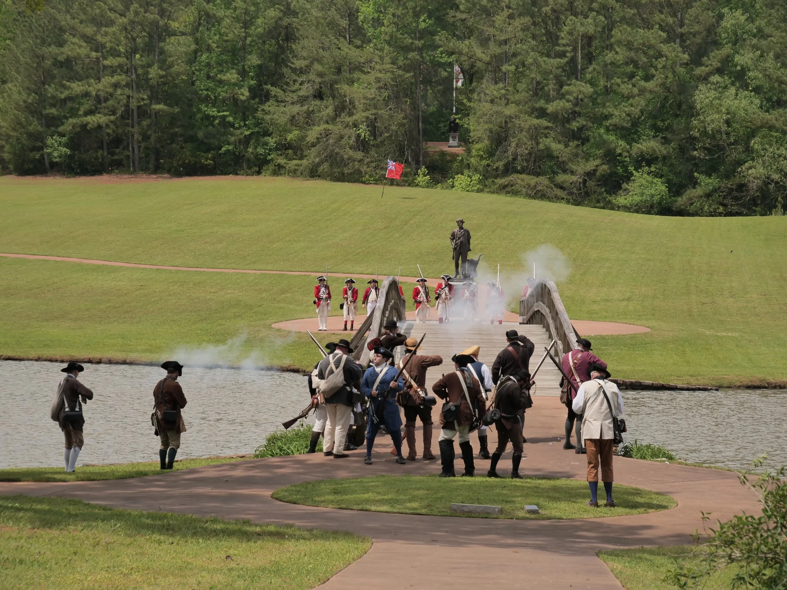 Reenactment of a historical battle at a park, featuring people dressed in colonial-era military uniforms, some with muskets, near a small lake, with a bridge and a statue of a man in the background, and smoke effects simulating gunfire.