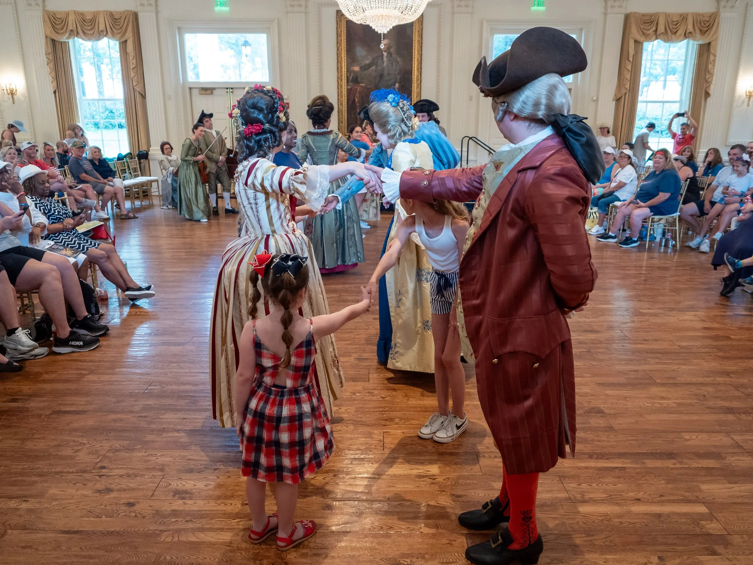 People dressed in 18th-century costumes dancing at a historical reenactment event in a large, elegant room with wood floors and large windows.