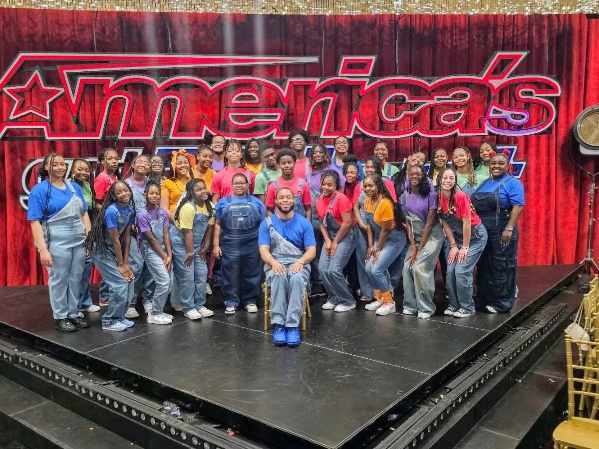 Birmingham Youth & Young Adult Fellowship Choir sitting and standing on a stage in front of a red curtain with a sign that reads 'America's Got Talent'. The group is smiling, wearing colorful t-shirts and overalls, and posing for a group photo.