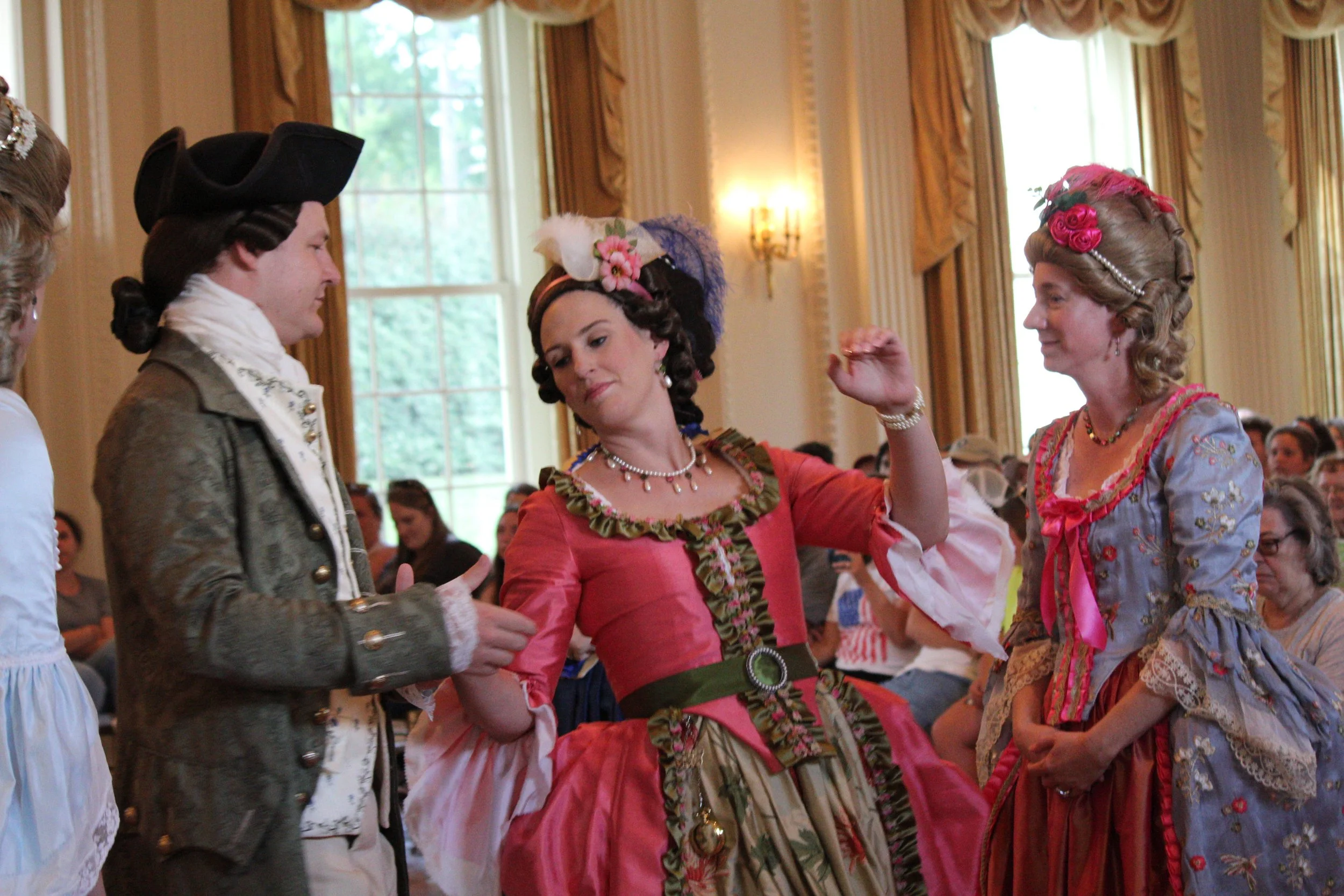 People dressed in 18th-century European aristocratic costumes, including a man in a tricorn hat and women in elaborate dresses, engaging in a social dance or gathering inside an elegant room with large windows and ornate decor.